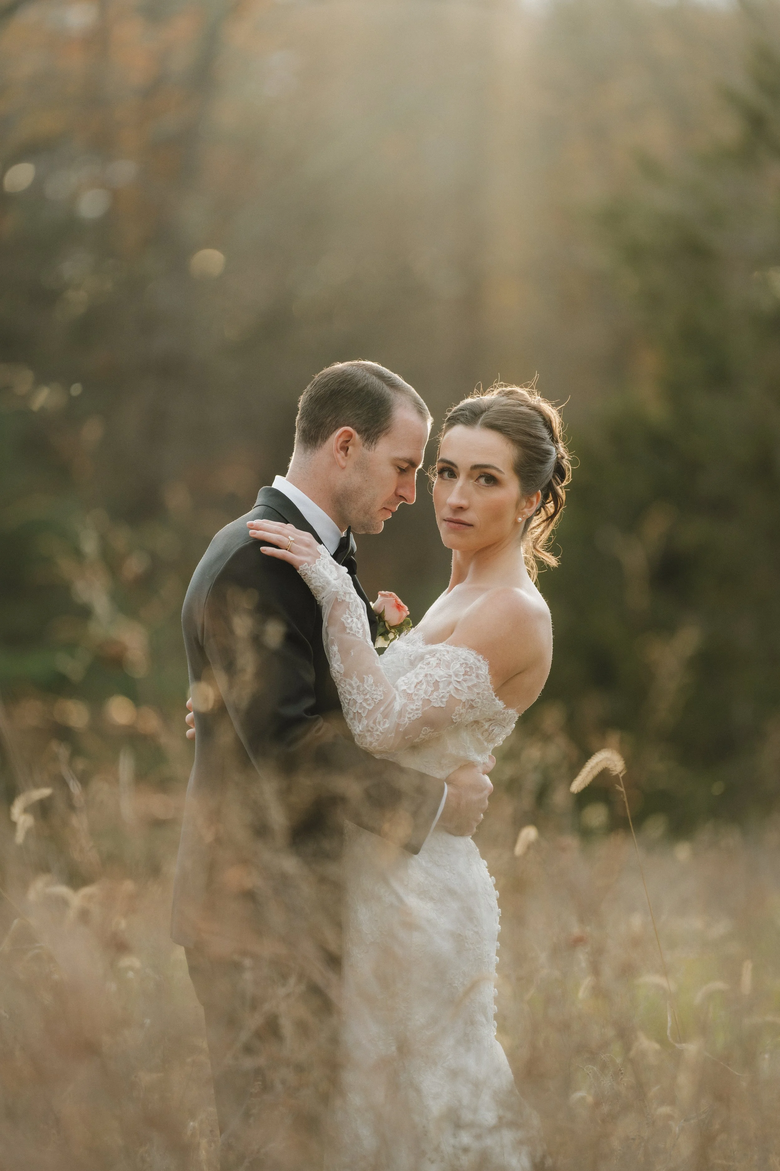A bride and groom standing close together in a natural outdoor setting during sunset, with the bride wearing a lace wedding gown and the groom in a black tuxedo.