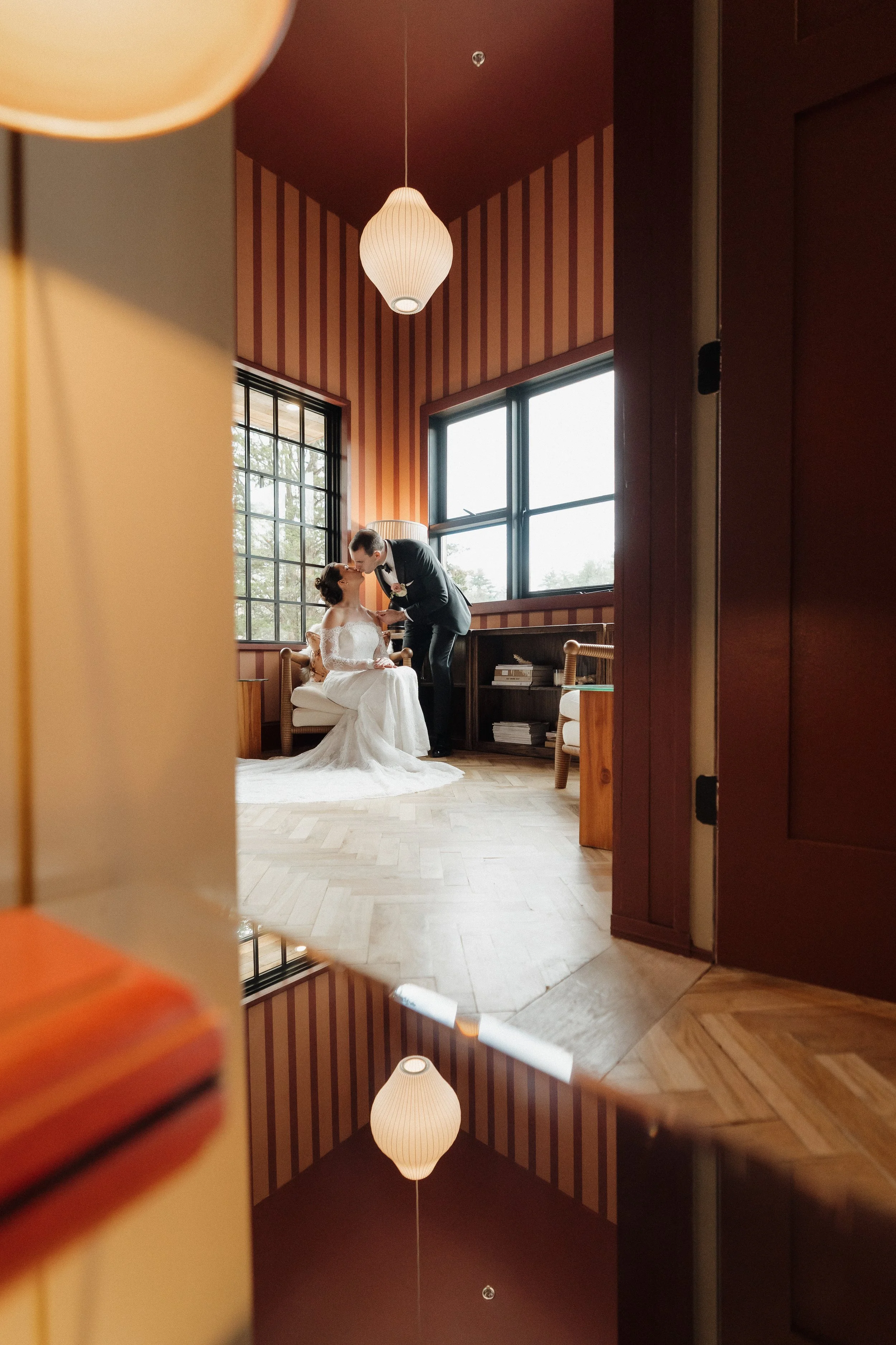 A bride and groom sharing a moment indoors, with the groom leaning in towards the bride who is seated on a chair, surrounded by wooden furniture and striped wallpaper, reflected in a mirror.