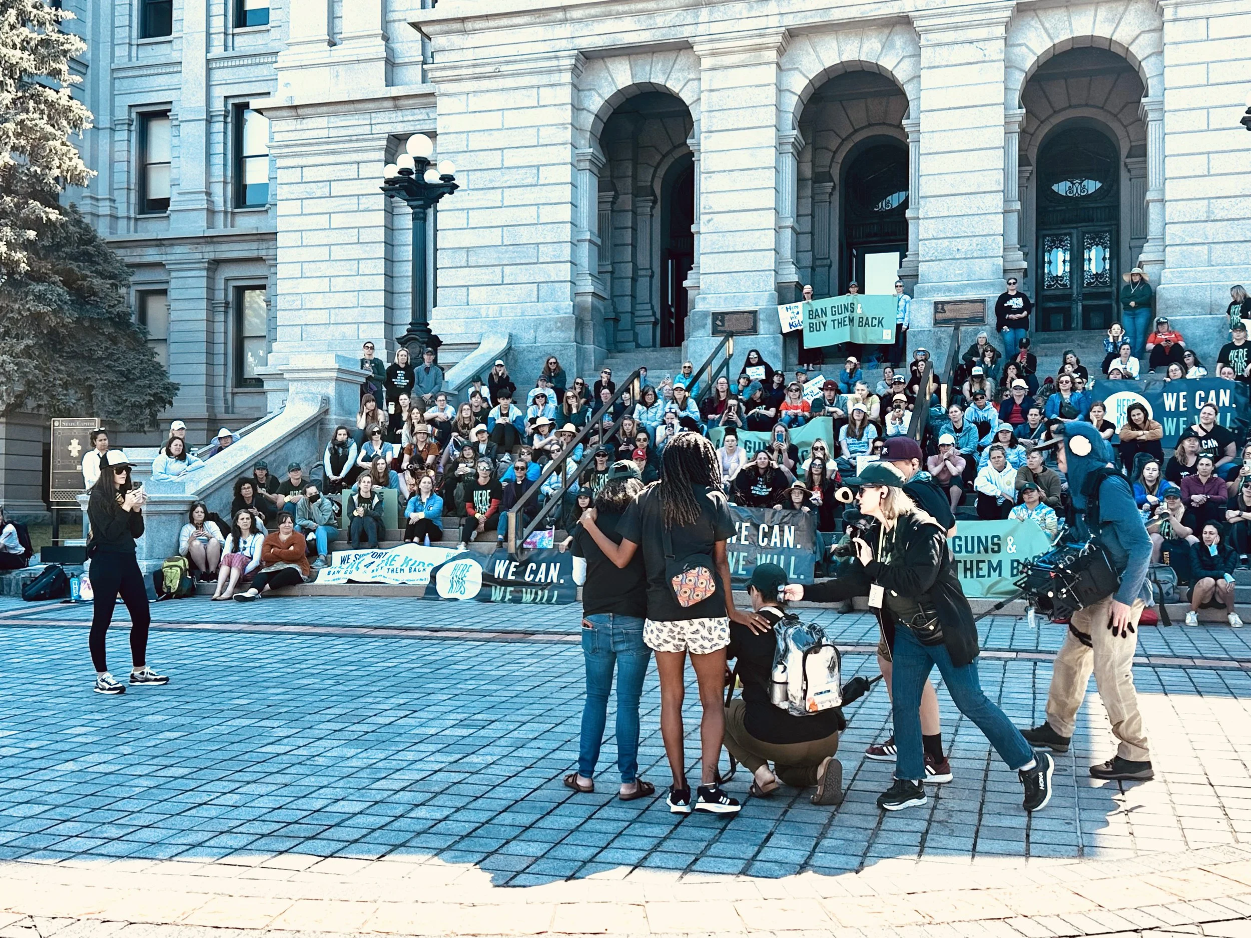 Image of a brown woman and a black woman facing a crowd of women sitting on the steps of the Denver capitol.