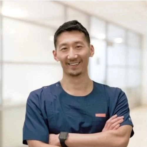 A smiling man in blue medical scrubs standing with arms crossed in a clinical setting.