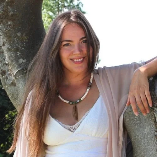 A woman with long brown hair smiling outdoors, leaning against a tree, wearing a white top and a beaded necklace.