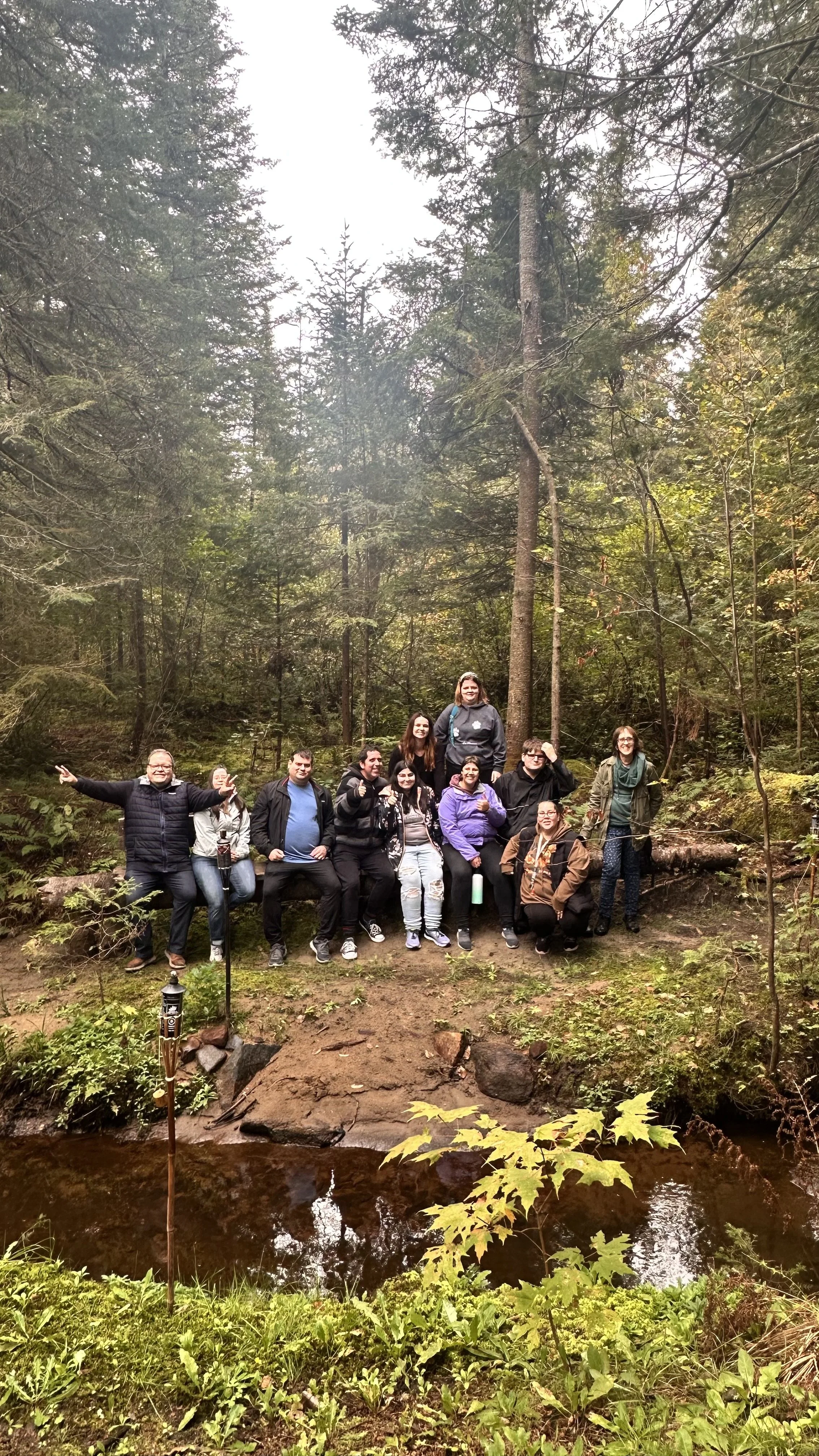 Un groupe de dix personnes posant sur un troncs au bord d'une petite rivière dans une forêt dense, entourée d'arbres verts.