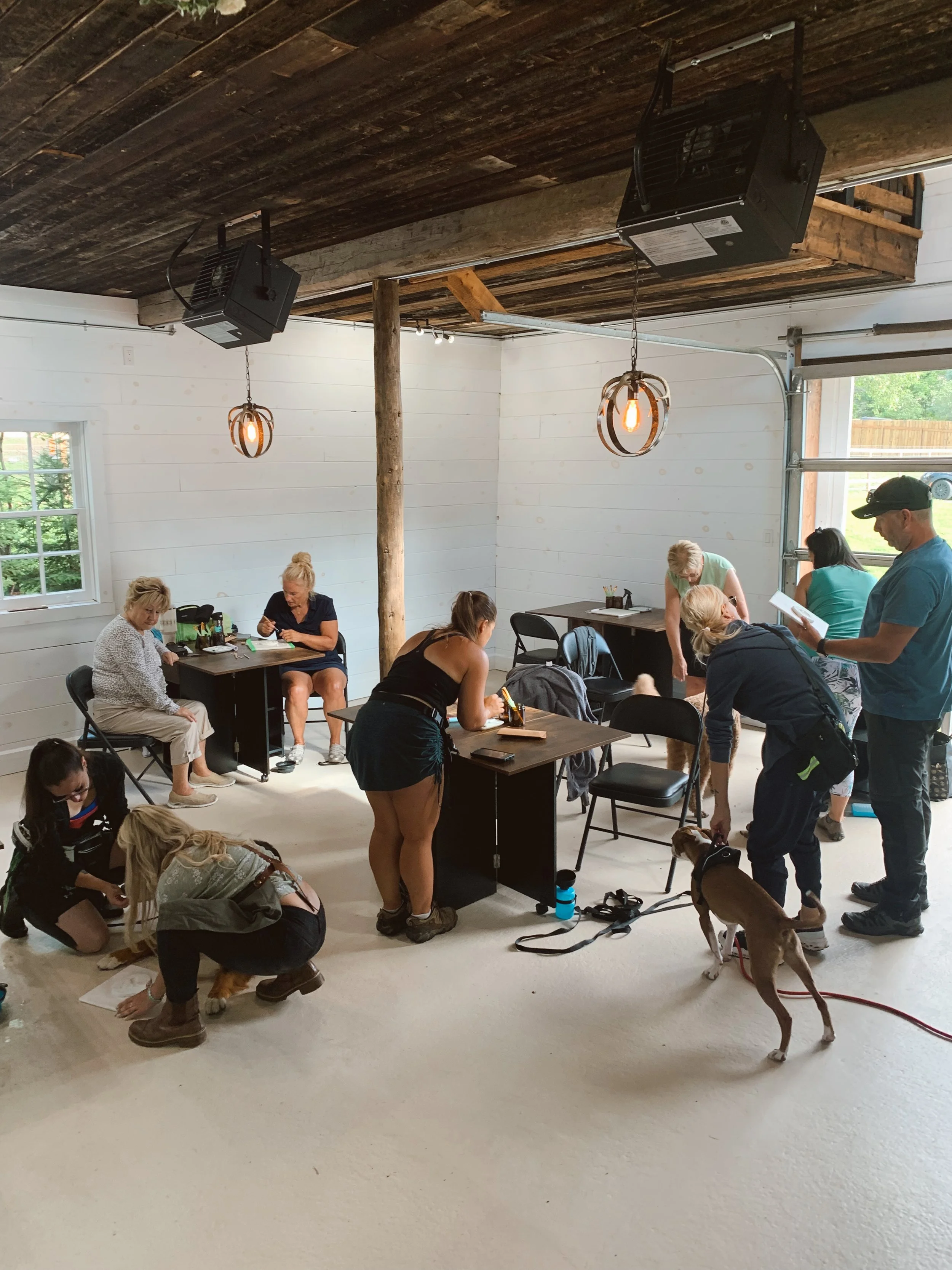 Une salle avec des personnes assises, discutant et s'occupant de chiens lors d'une session ou atelier, avec un intérieur en bois et une grande porte de garage ouverte.