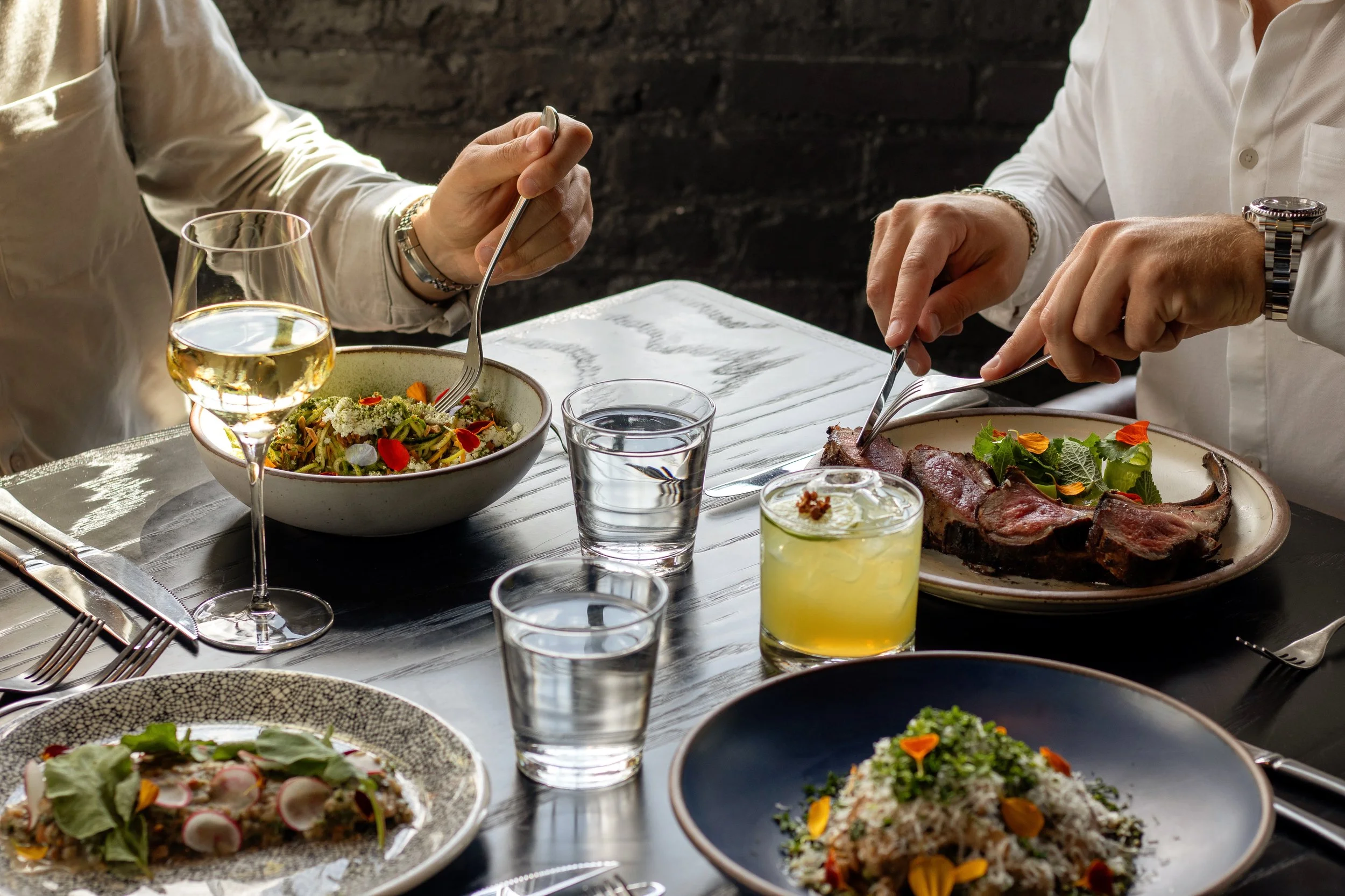 Close-up of a dining table with various dishes, including a bowl of salad, a plate of cooked meat, and glasses of wine and water, with two people serving food.