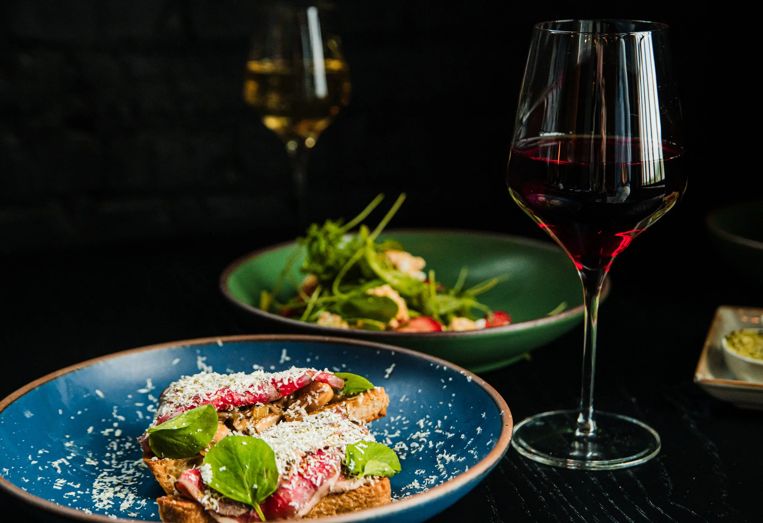 Close-up of a plate with a sandwich topped with cheese, slices of radish, and greens. In the background, a glass of red wine and a glass of white wine are visible, with a dark table setting and a blurred ambiance.