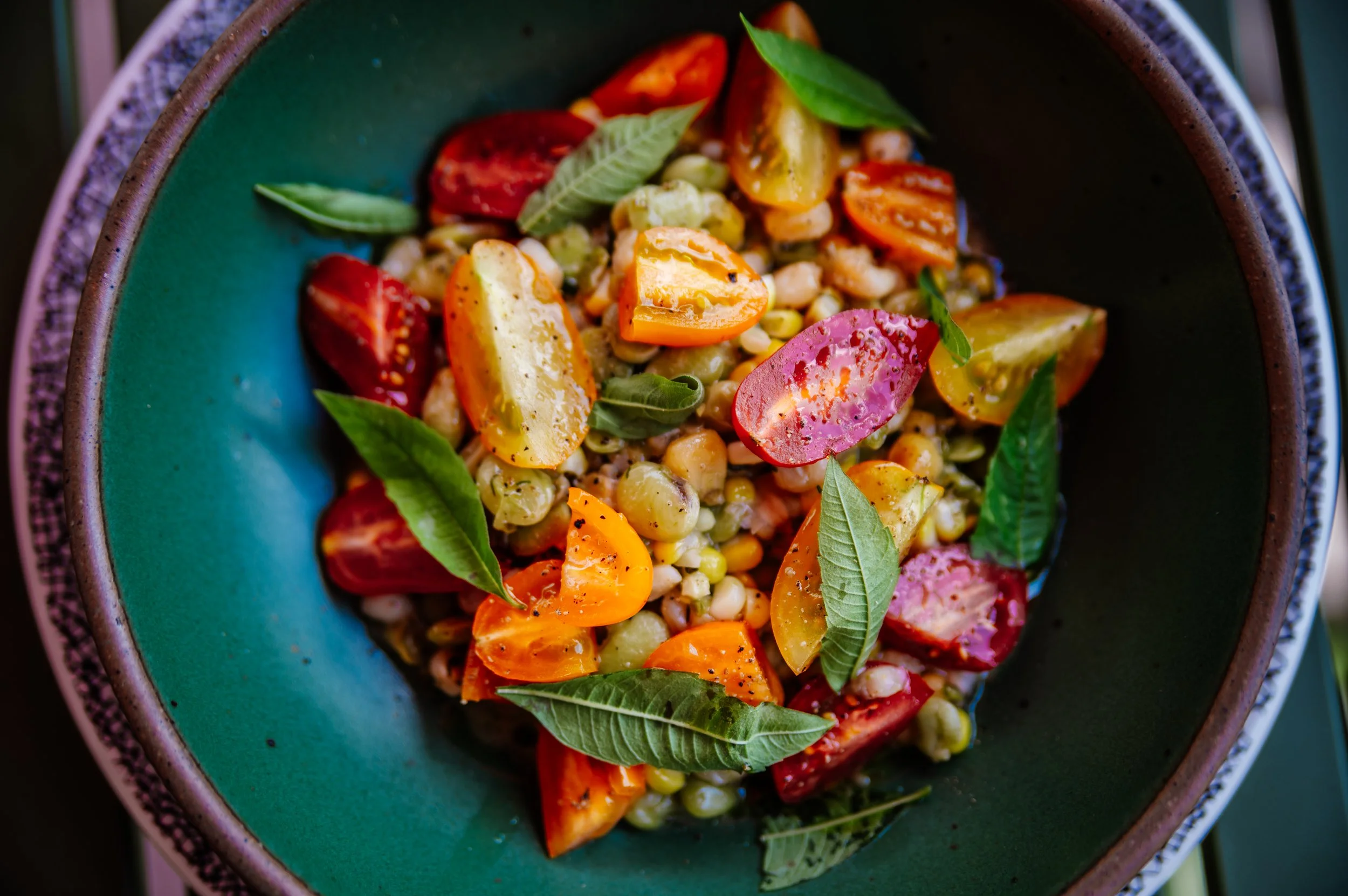 Close-up of a bowl containing a colorful dish with cherry tomatoes, corn, fresh basil leaves, and possibly grilled vegetables.