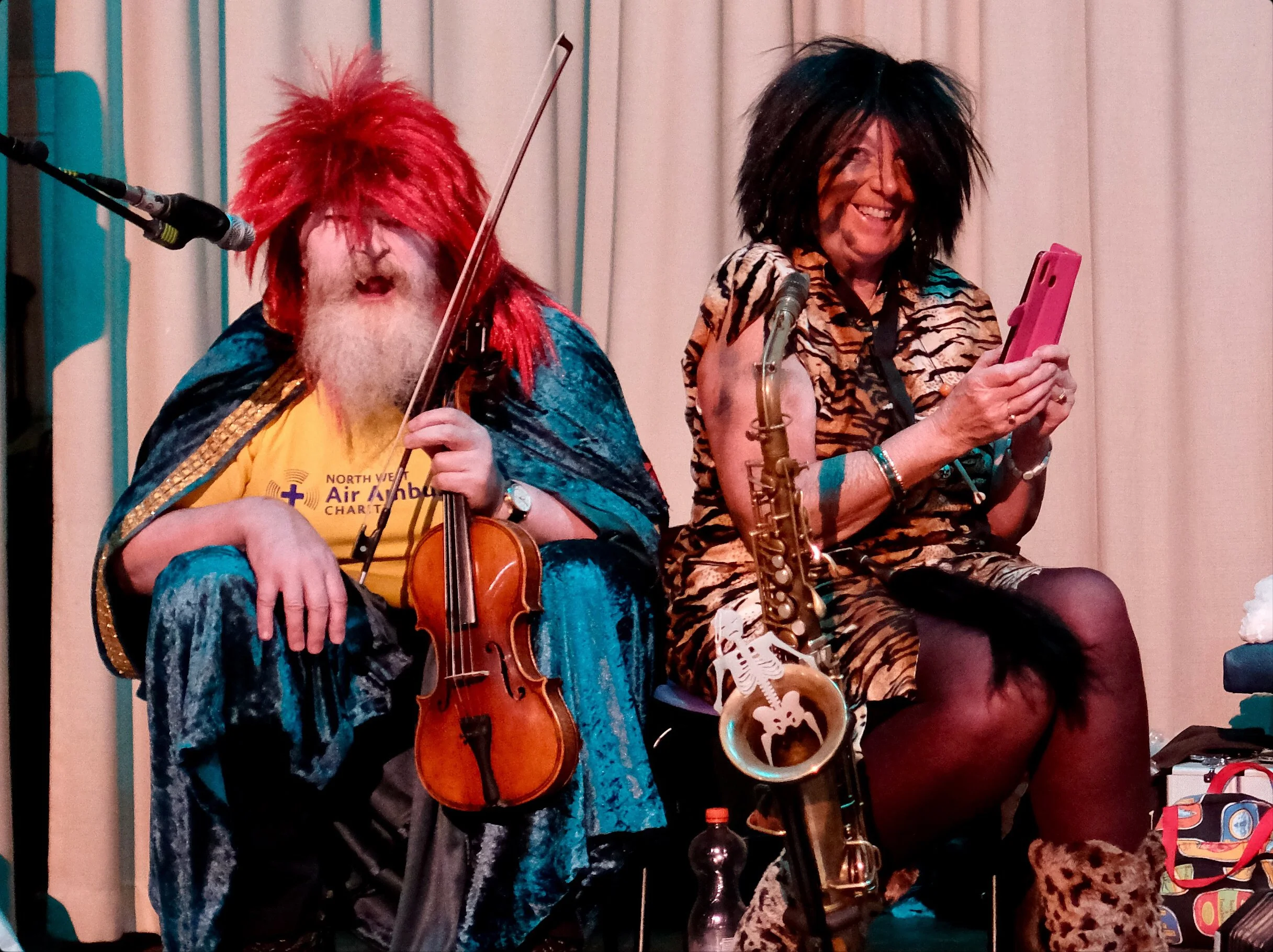 Two performers dressed in colorful, eccentric costumes; one with a red wig and blazer, playing the violin, and the other with a black wig, animal print dress, and floral boots, holding a pink smartphone, on a stage with a curtain backdrop.