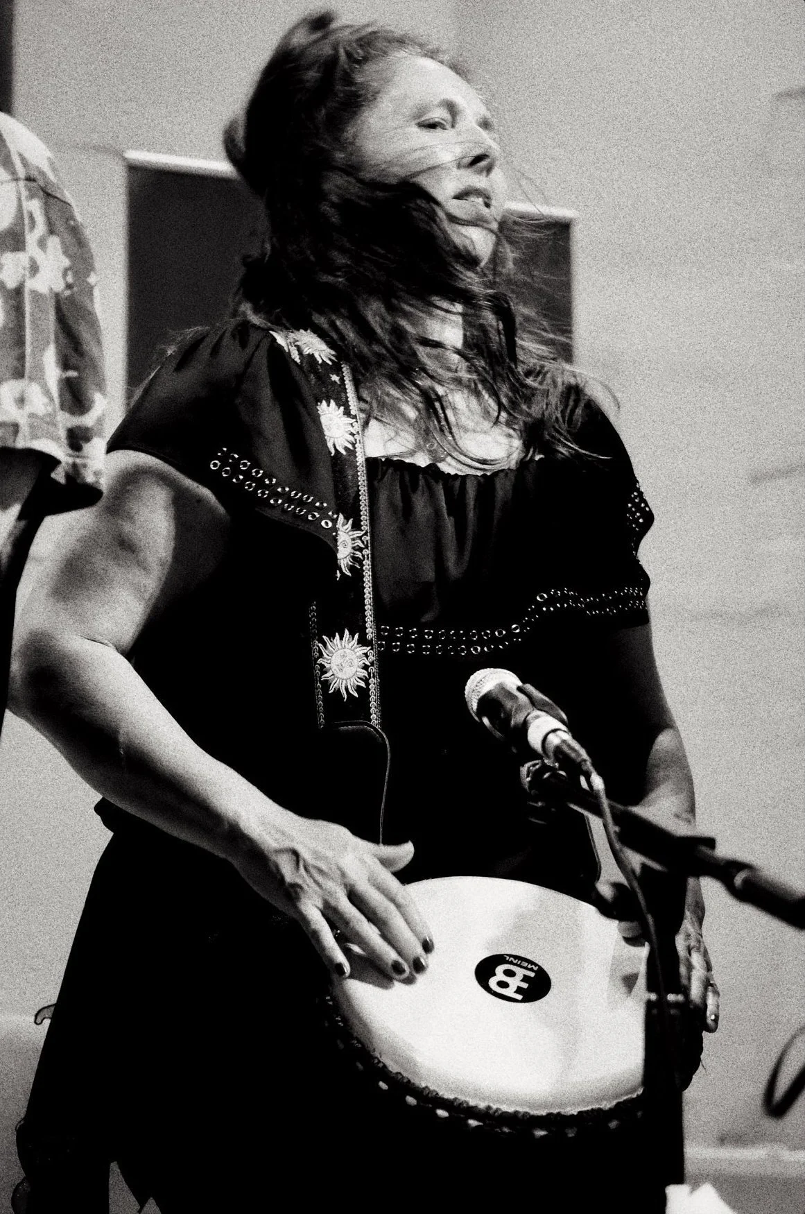 A woman with long hair playing a hand drum at a microphone in a black and white photo.
