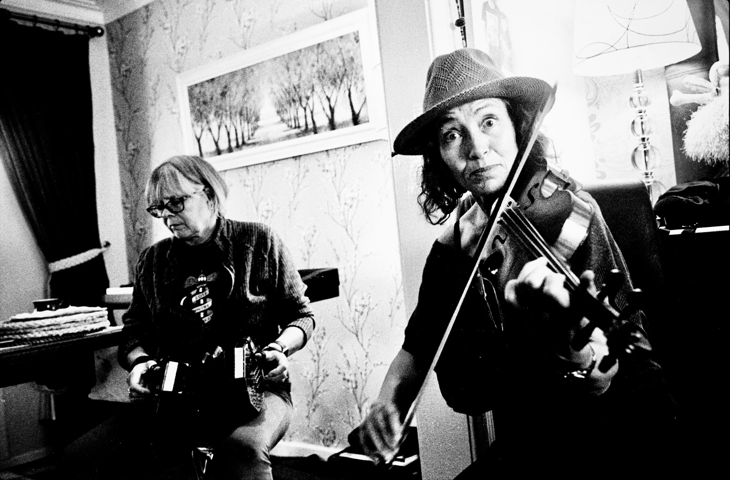 Two women playing musical instruments in a room, one with a violin and the other with an accordion, with artwork on the wall behind them.