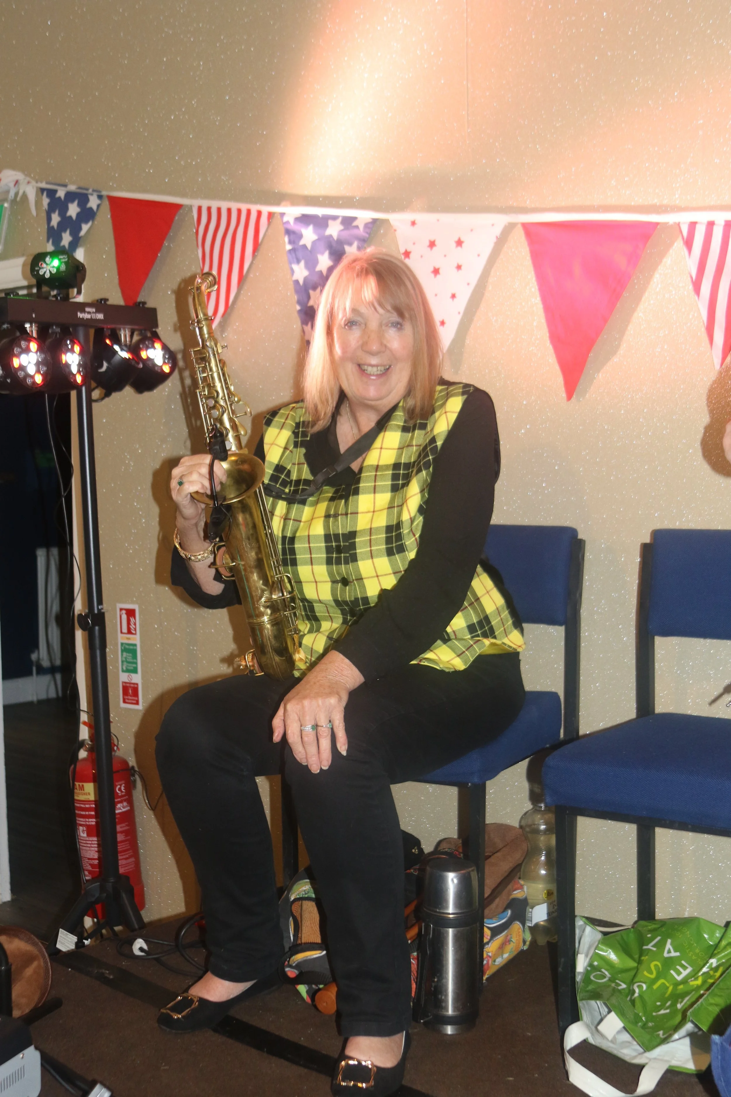 A woman sitting on a blue chair holding a saxophone, smiling with festive bunting in the background.