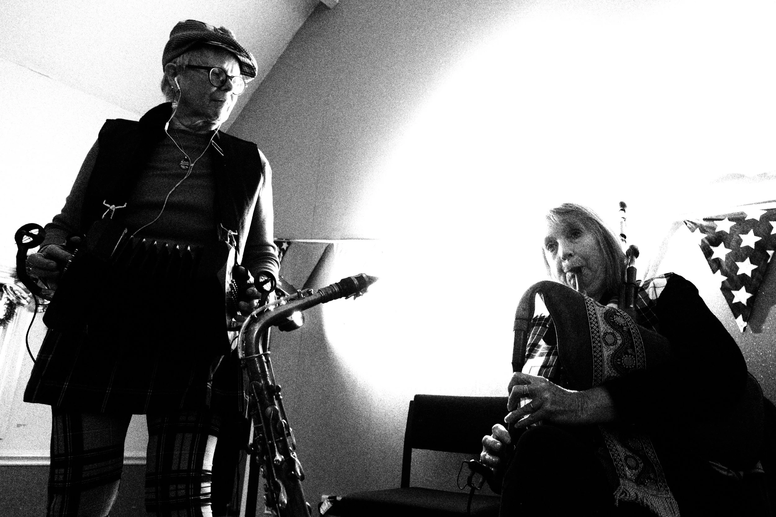 Two women playing musical instruments indoors; one woman with glasses and a headscarf is playing a saxophone, and another woman is playing a clarinet or similar wind instrument; patriotic star decorations in the background.