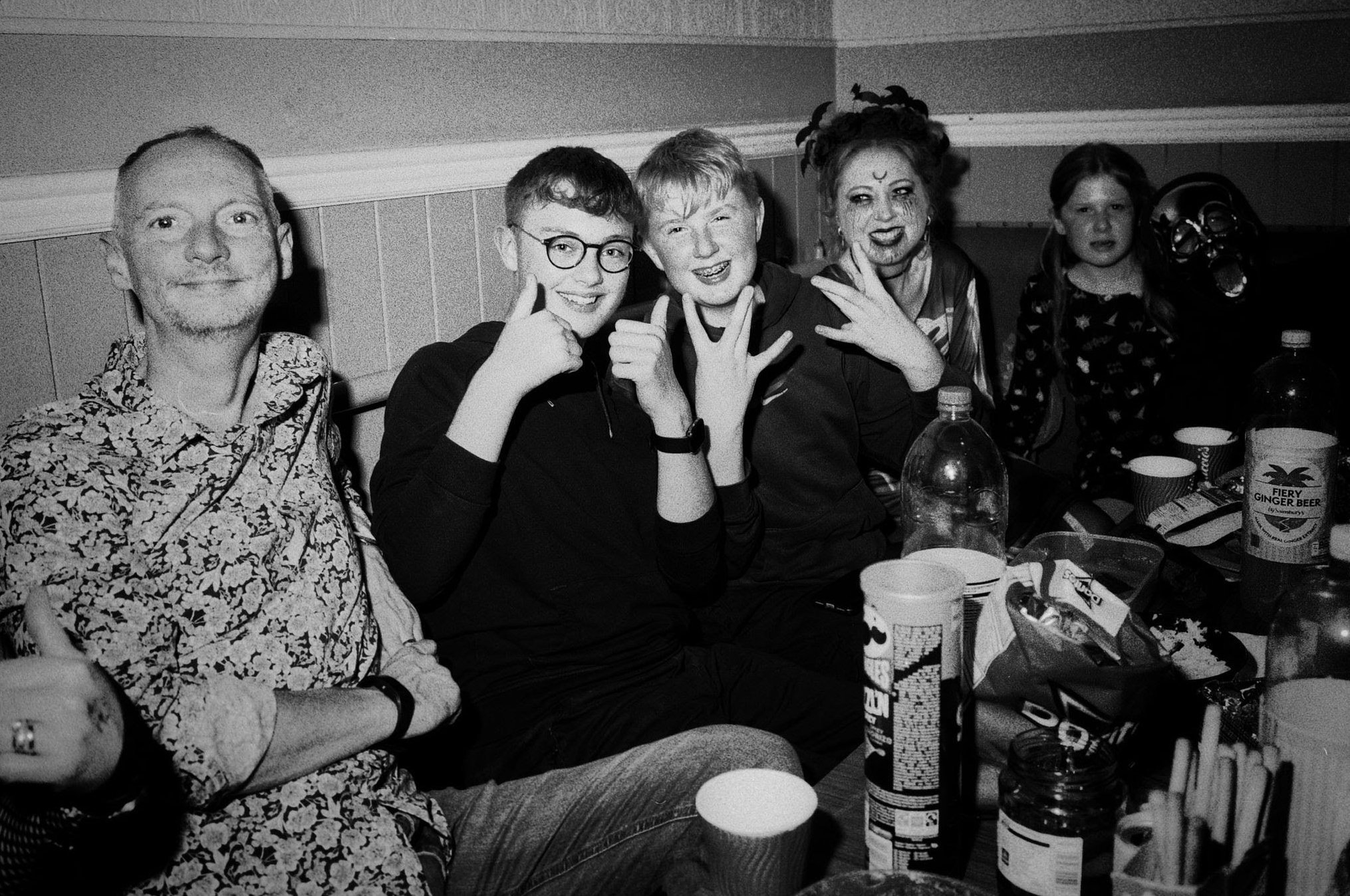 Group of six people sitting at a table during a gathering or party, with snacks and drinks, smiling and posing for the photo. One person is wearing a mask.