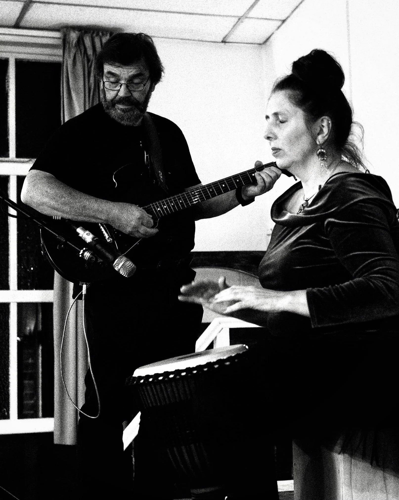 A man playing guitar and a woman singing and playing a instrument in a room with window curtains.