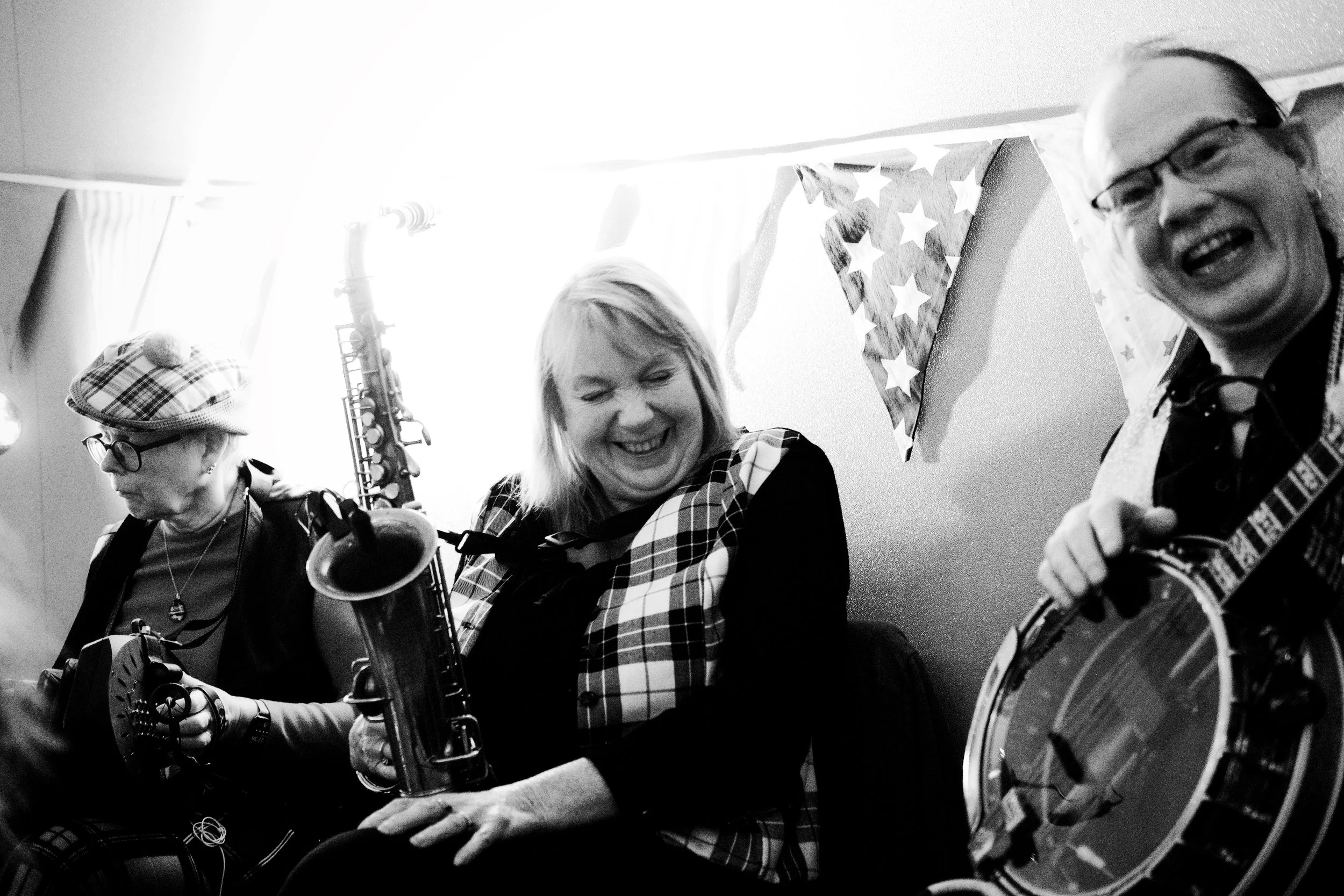 Three people playing musical instruments at a celebration with patriotic bunting. The person on the left is wearing glasses and a hat, playing a guitar. The person in the middle is a woman laughing, wearing a plaid vest, holding a saxophone. The pers