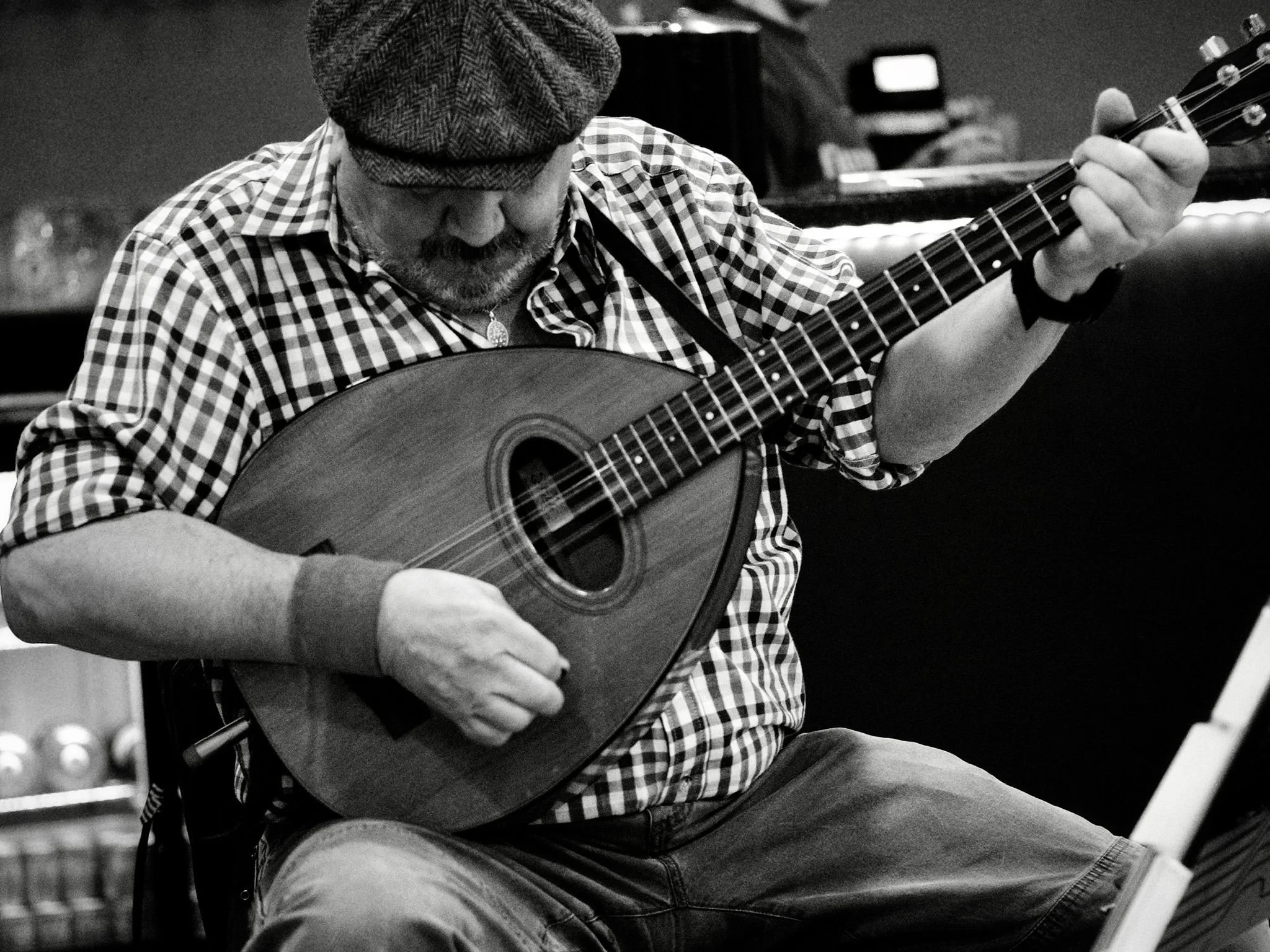 A man wearing a checkered shirt, a hat, and glasses playing a mandolin.