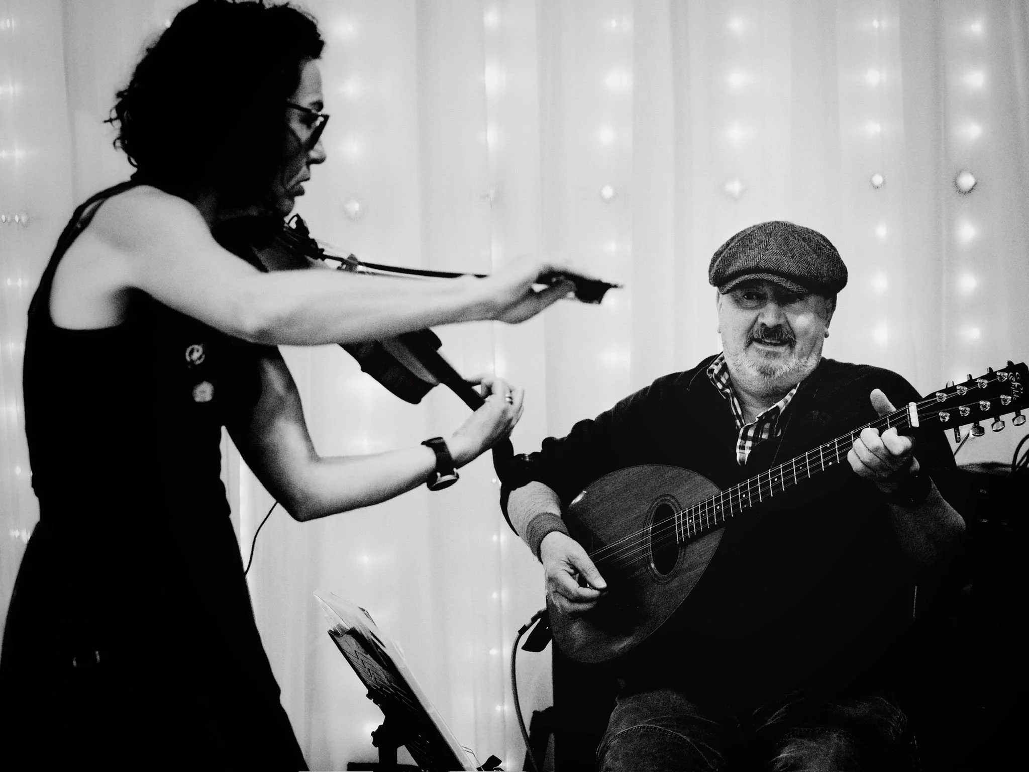A black and white photo of a woman playing the violin and a man playing the guitar, with the woman standing and the man seated, in a setting with string lights in the background.