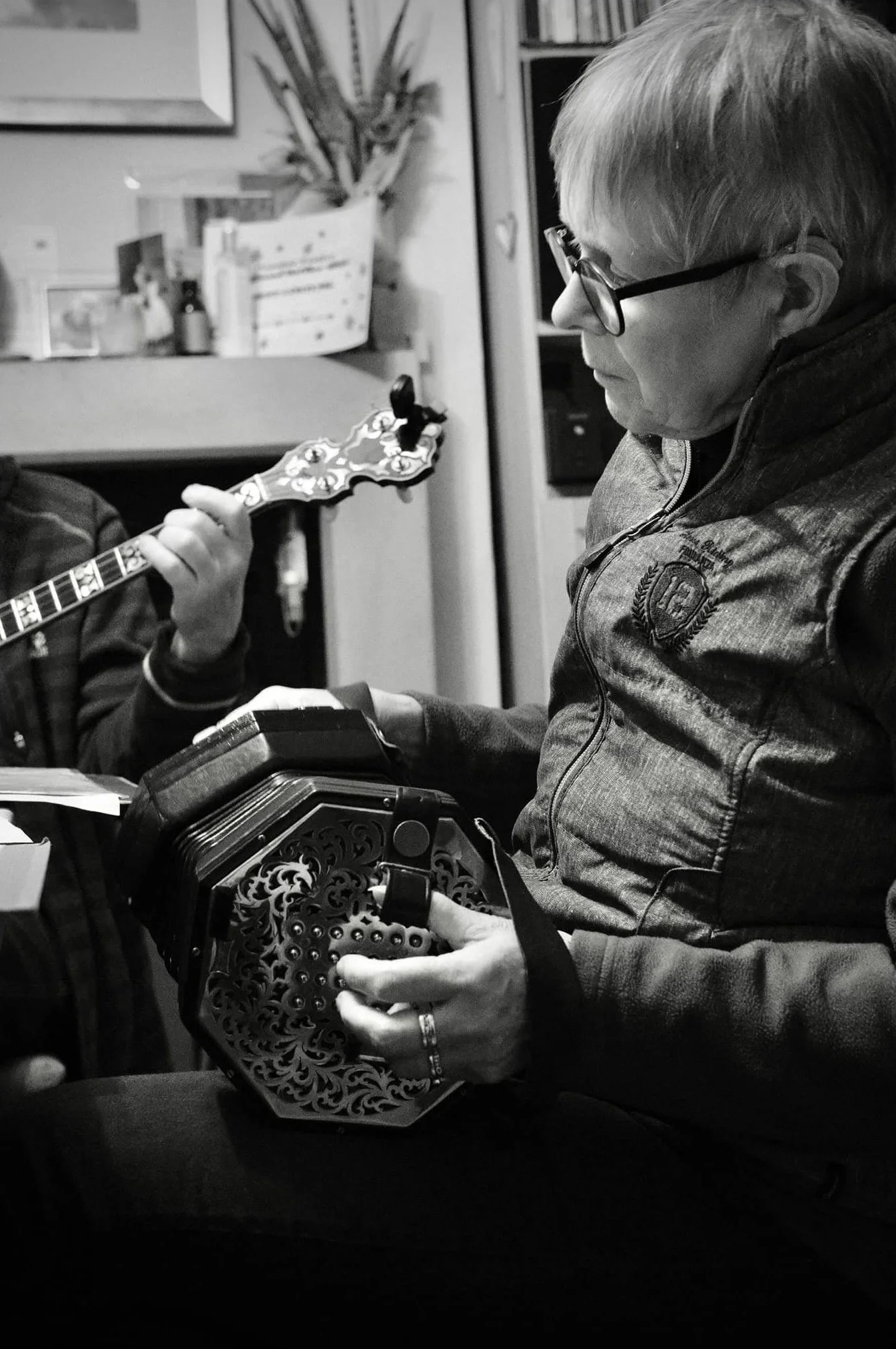 A woman sitting indoors, holding a melodica, with a person behind her playing a guitar.