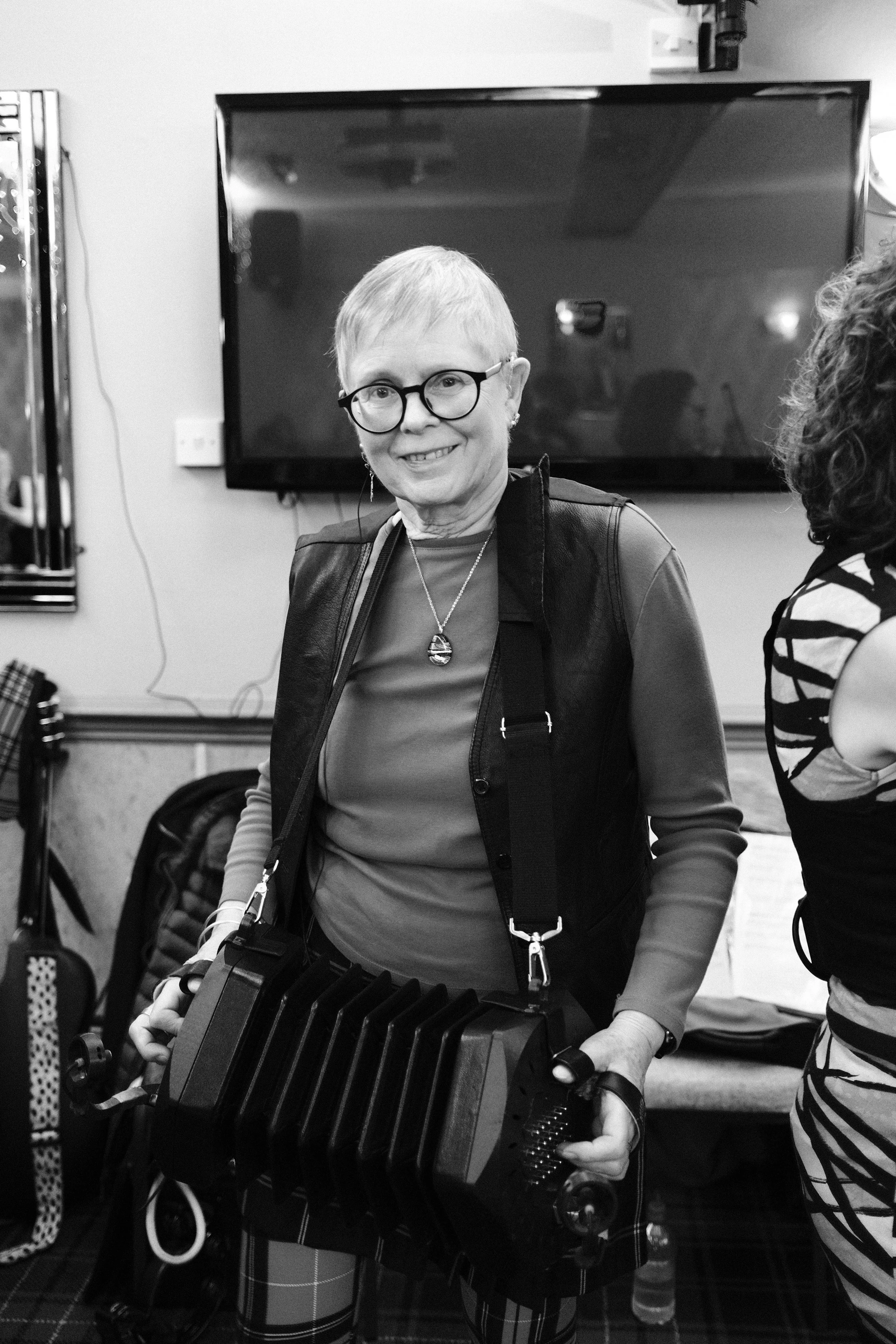 A smiling woman with short hair, glasses, and earrings, wearing a leather vest over a long sleeve shirt, holding a large accordion in a room with a TV on the wall behind her.