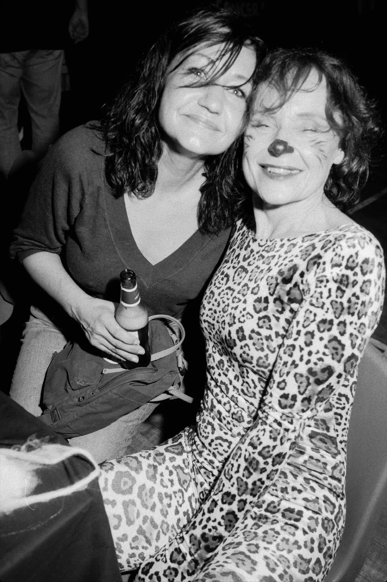 Two women at a social event, one holding a beer bottle, wearing casual clothes including a leopard print outfit and a dark V-neck top.
