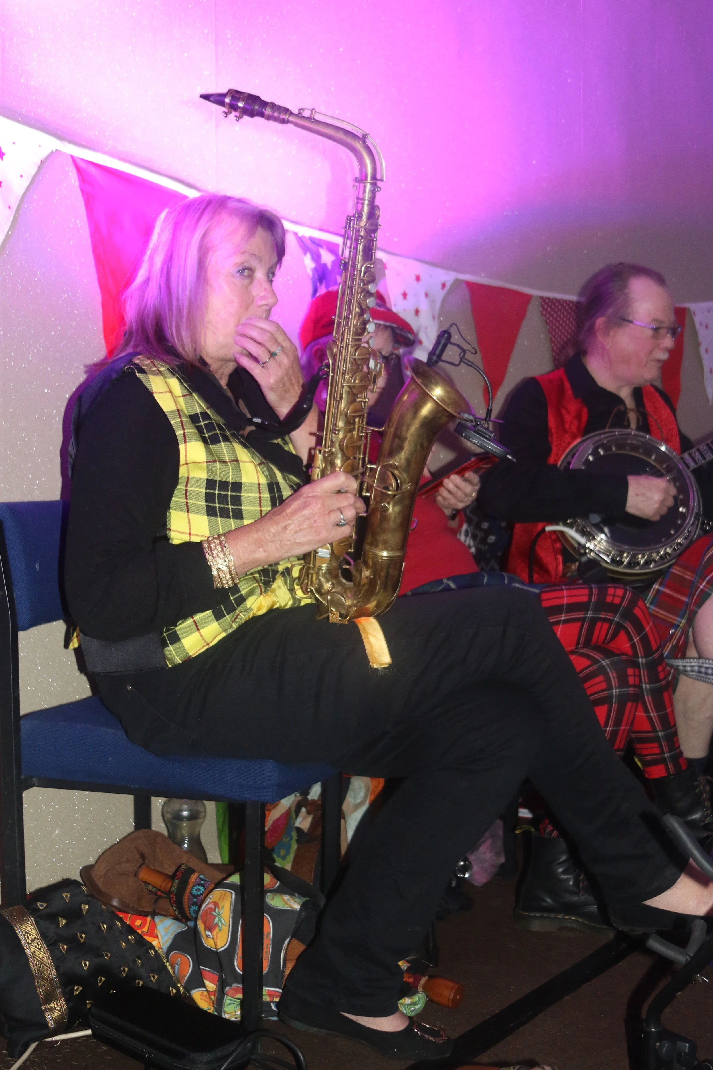 A woman playing a saxophone at a festive event with women in red and black plaid clothing, bunting flags on the wall, under pink lighting.