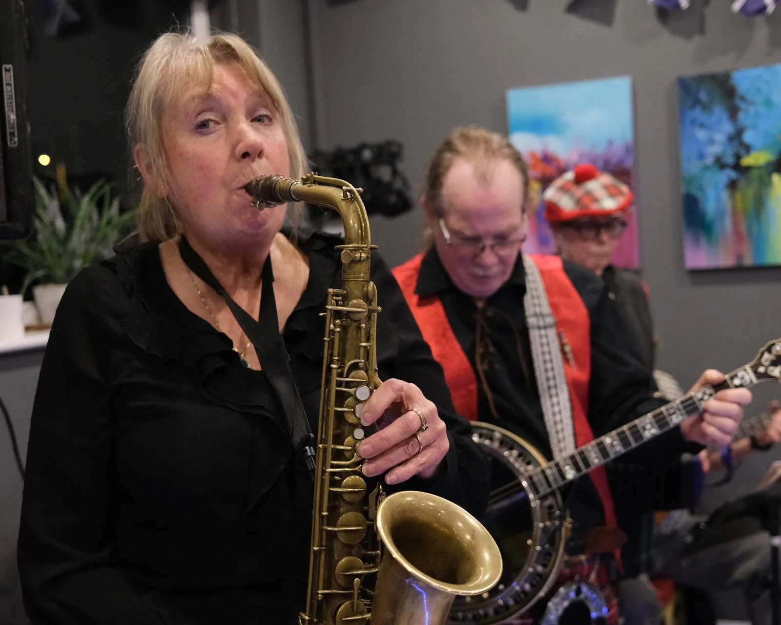 Three musicians performing indoors, with the woman in the foreground playing a saxophone, and two men in the background playing banjos.