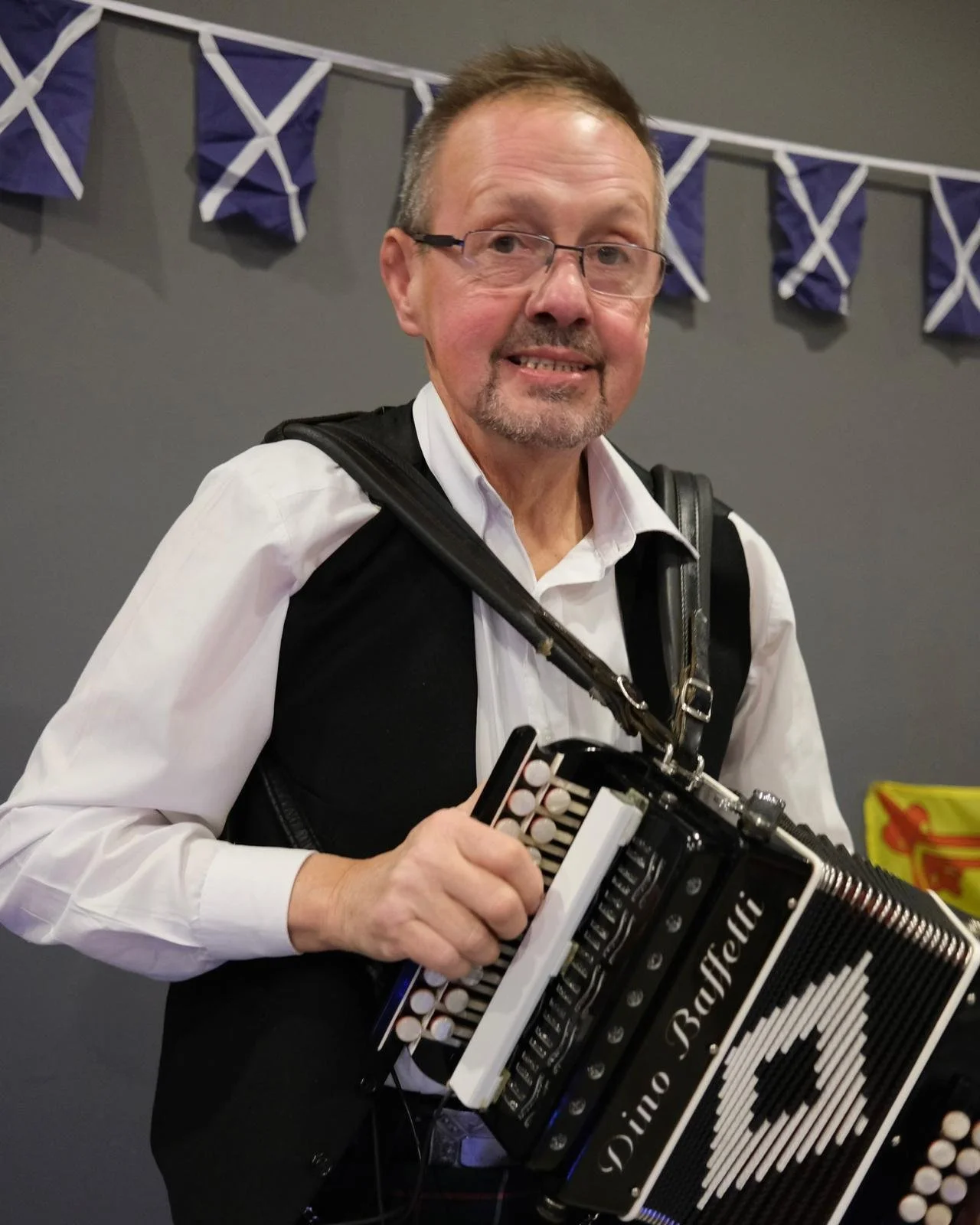A man with glasses, wearing a white shirt and a black vest, playing a black and white diatonic accordion.