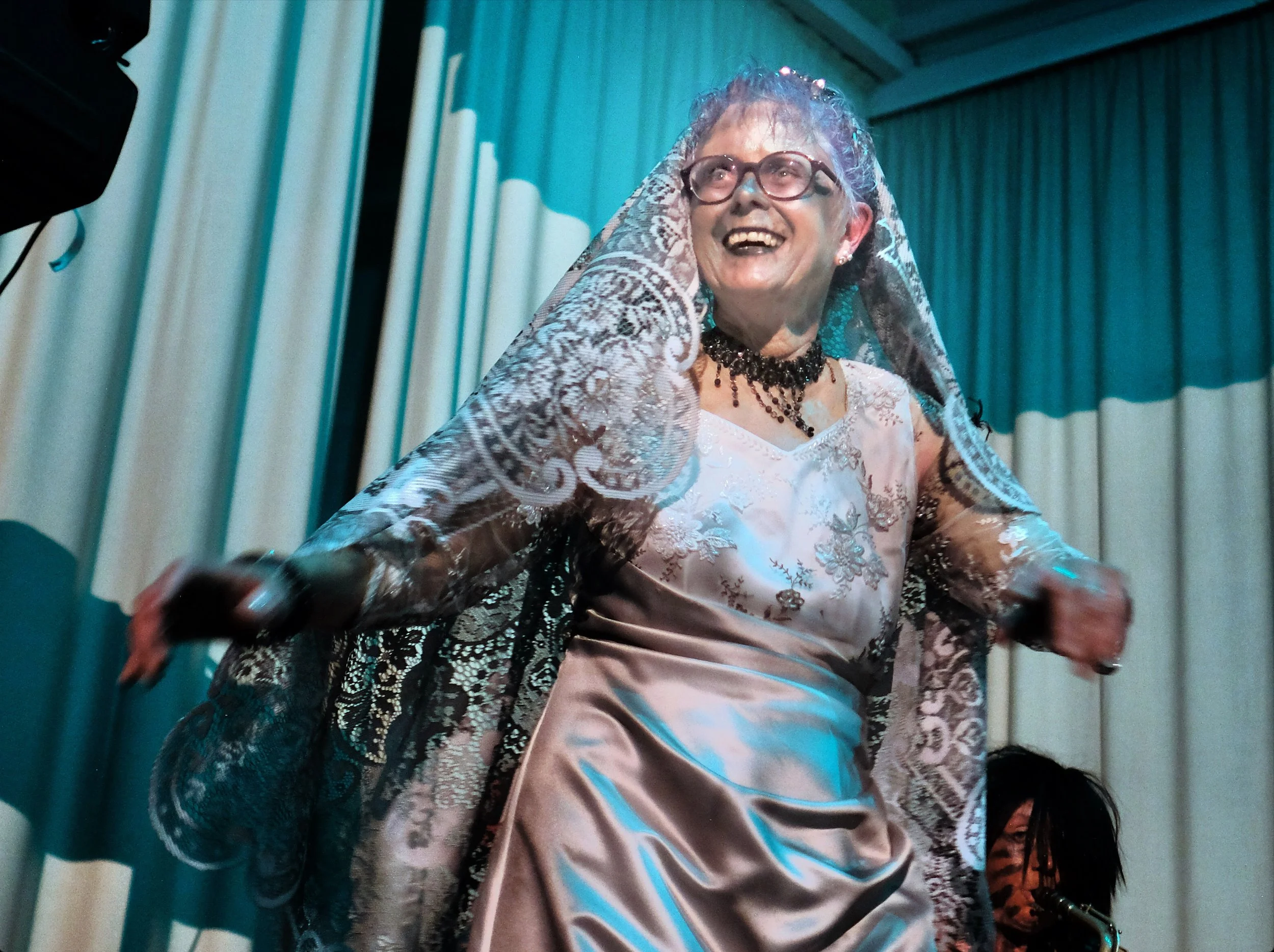 An elderly woman in a fancy dress smiling and laughing, wearing glasses and a veil, on a stage with curtains behind her.