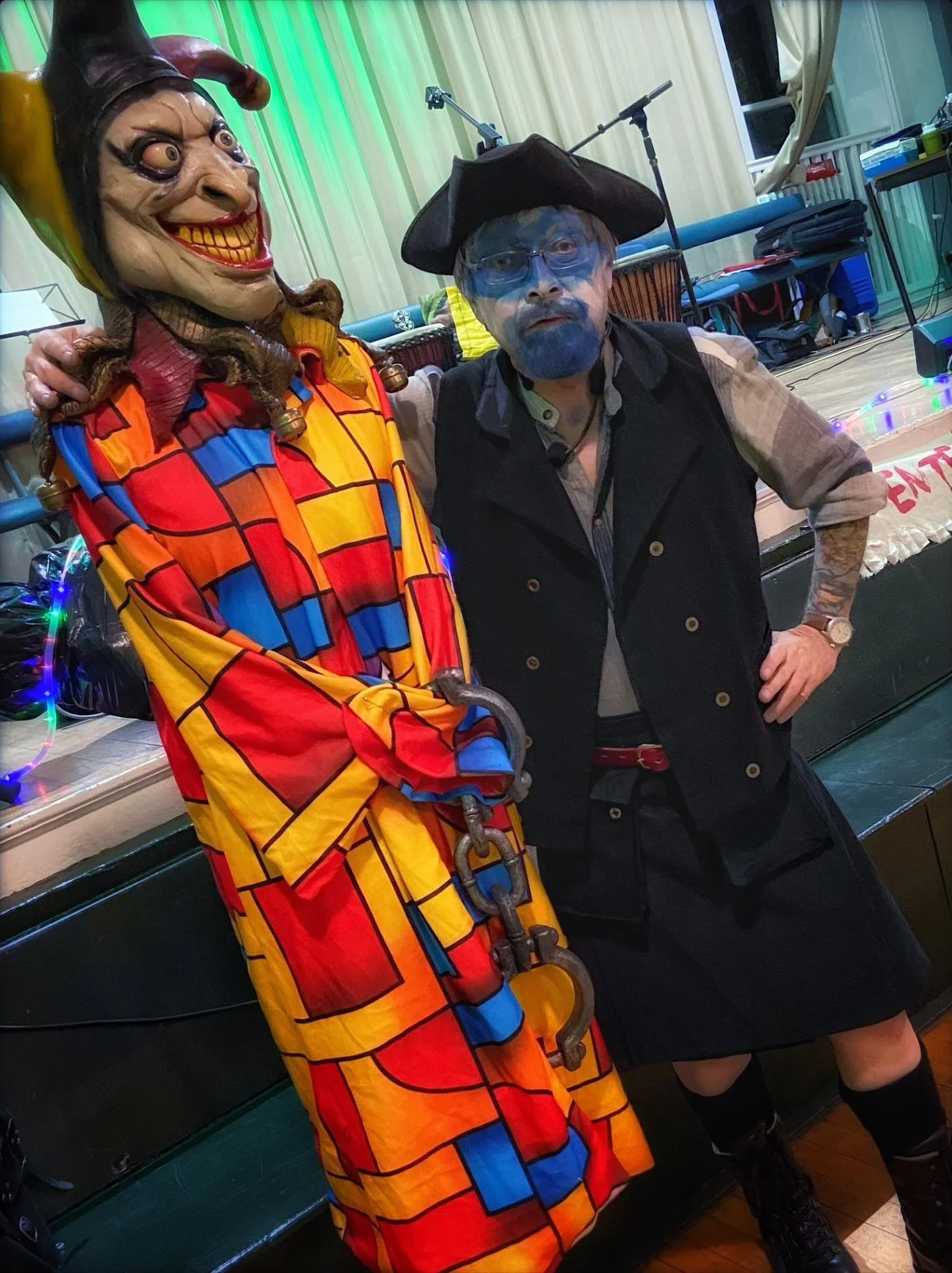 A person dressed as a clown with a large, smiling mask wearing a colorful, geometric-patterned robe stands next to a person dressed as a pirate with blue face paint, a pirate hat, and a vest in front of a stage with musical instruments and equipment.