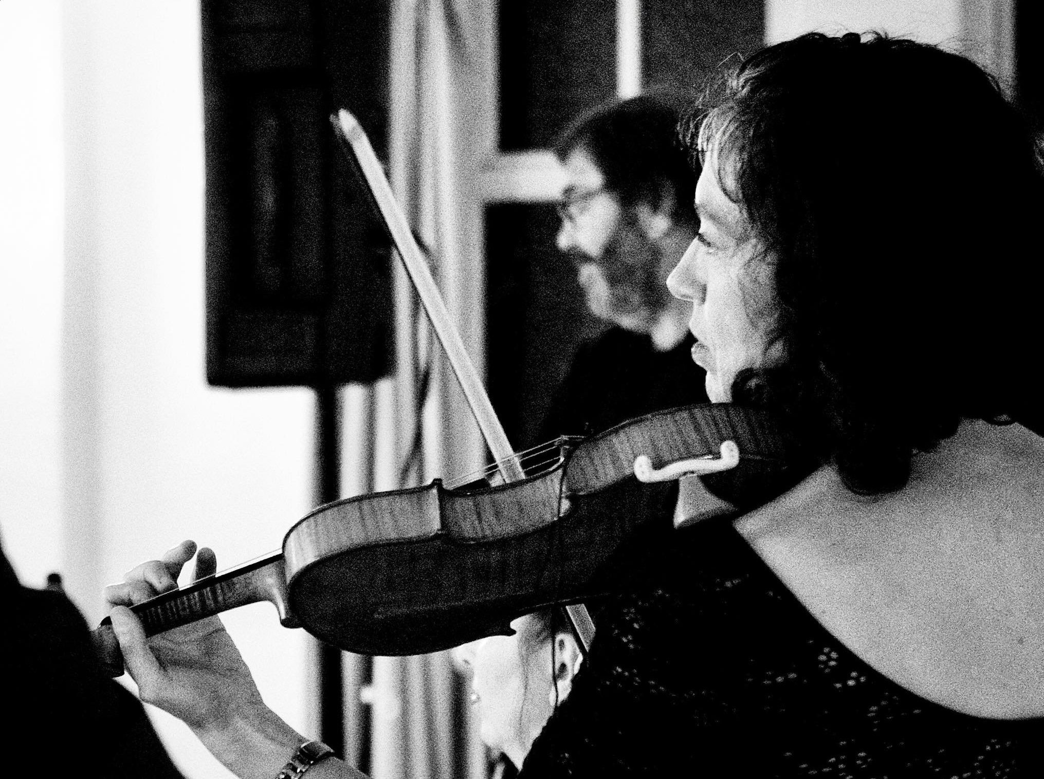 A woman playing the violin with a man in the background in a black and white photo.
