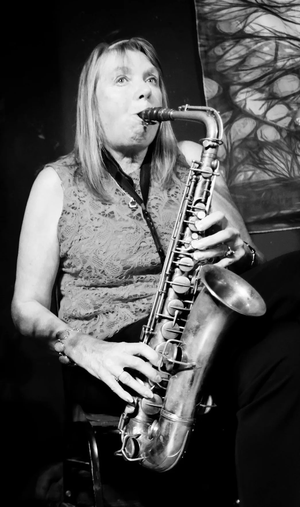 Black and white photo of a woman playing a saxophone, seated indoors against a background with abstract art.