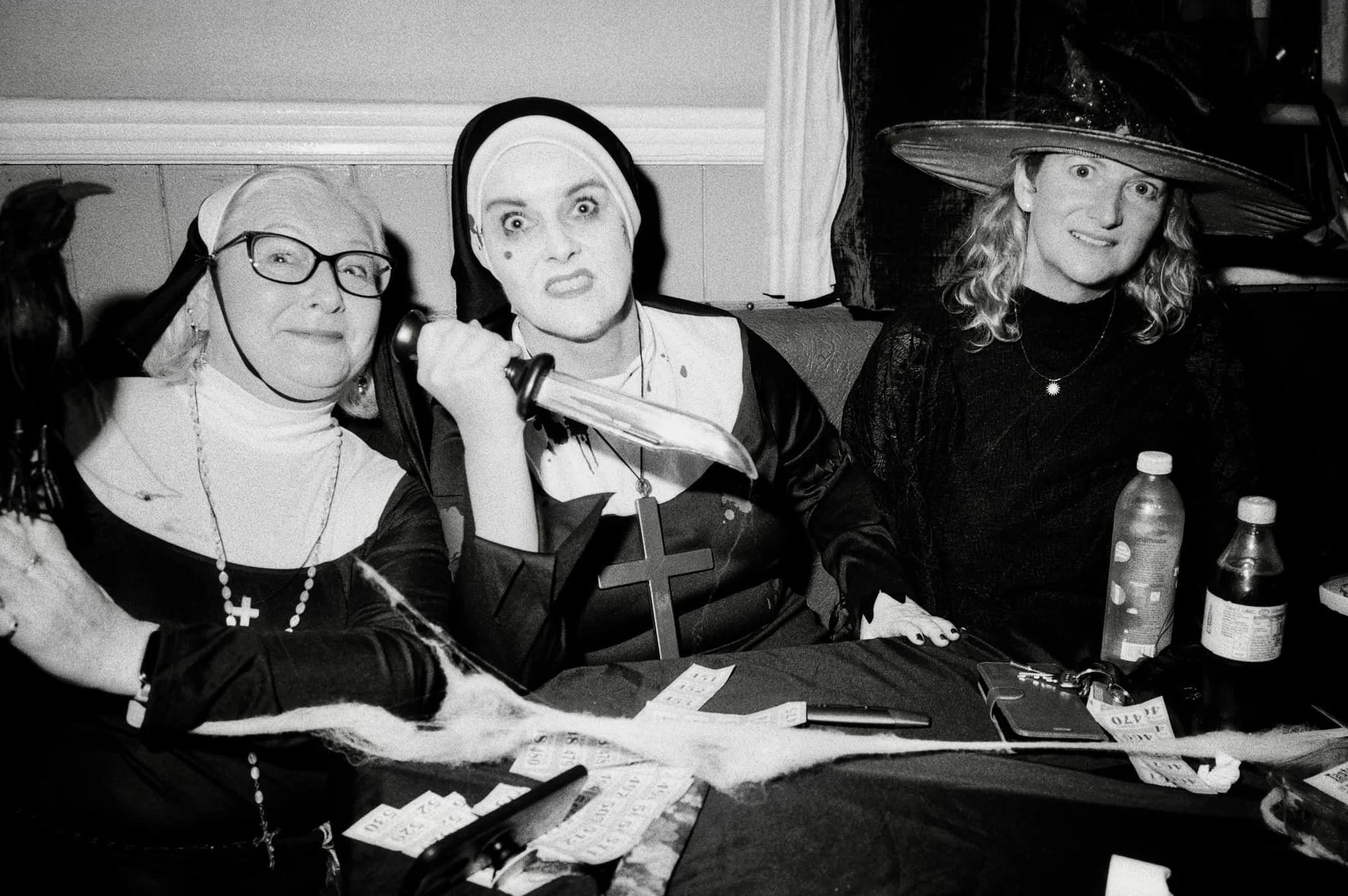 Three women dressed as witches sitting at a table with Halloween decorations, one holding a sword, another in a witch hat, all in costume for Halloween.