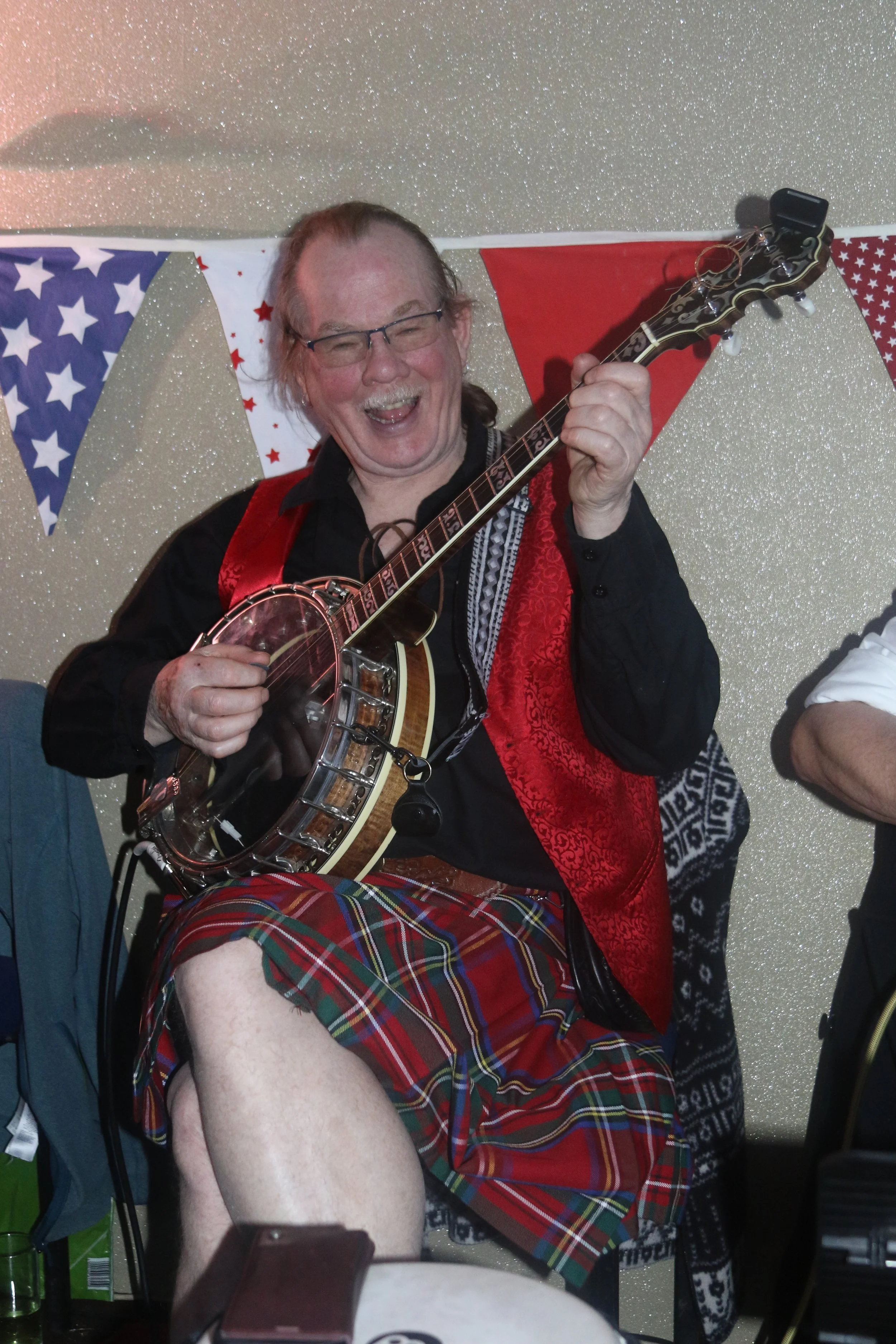 A man wearing glasses, a black shirt, red vest, and a tartan kilt, playing a banjo and smiling joyfully at a celebration with American flags and red, white, and blue decorations in the background.