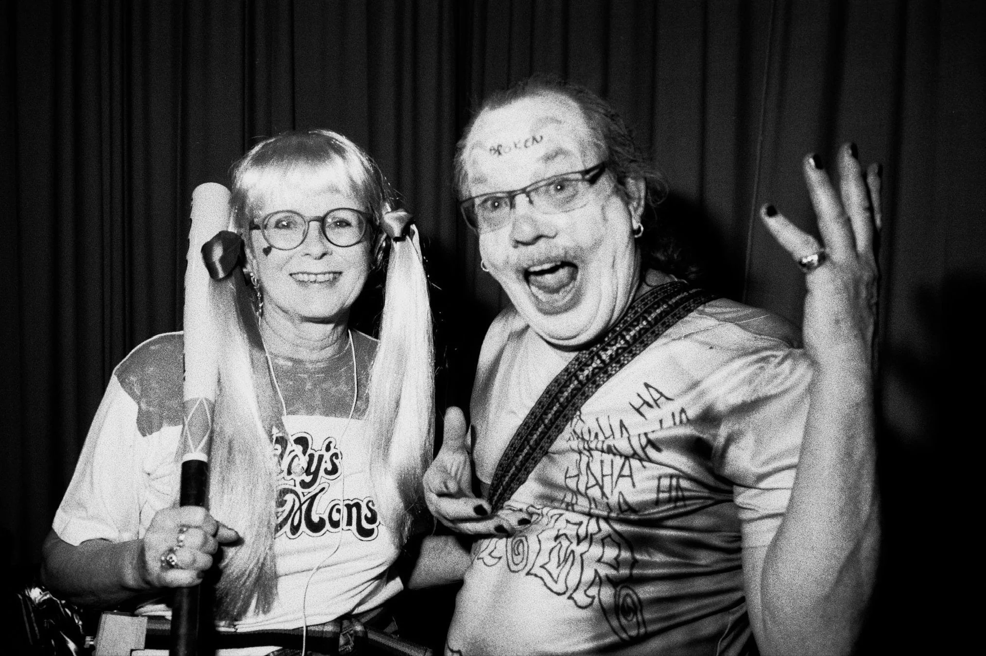 A woman with glasses and long hair styled in pigtails, wearing a 'Toys Dolls' shirt, holding a microphone, smiling. A man with glasses, shoulder-length hair, and a graphic shirt, has words written on his forehead, appears excited and is making a gest