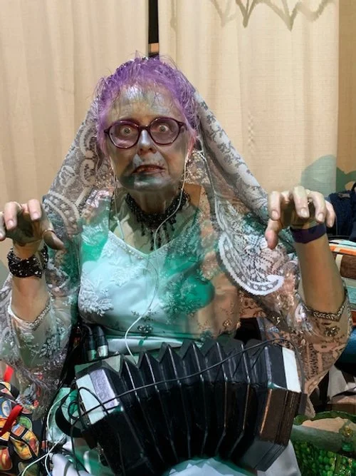 An older woman with purple curly hair, glasses, and a necklace, wearing a lace shawl and a green dress, holding her hands like claws in a playful, spooky pose, with a musical accordion in front of her.