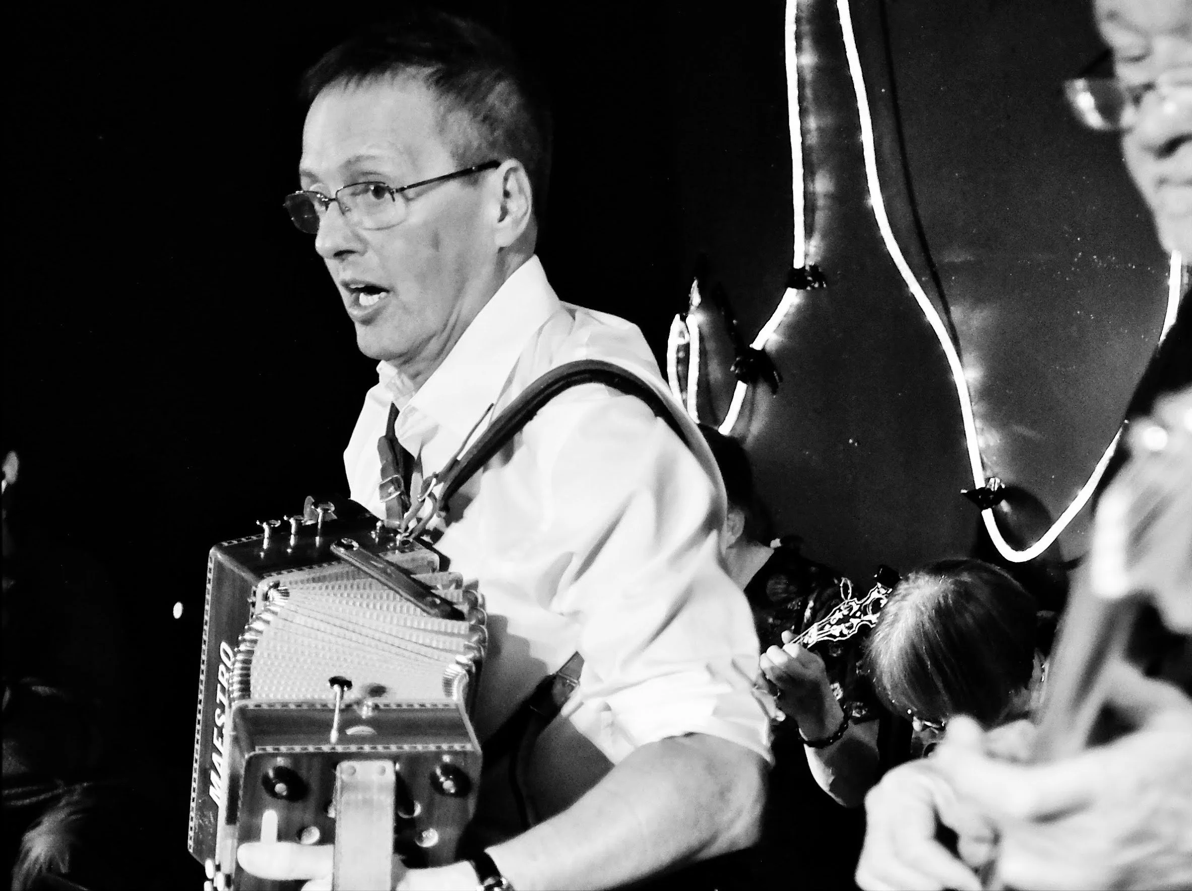 Black and white photo of a man wearing glasses and a white shirt, playing a musical instrument with a distinctive design, surrounded by other musicians, with a dark background.