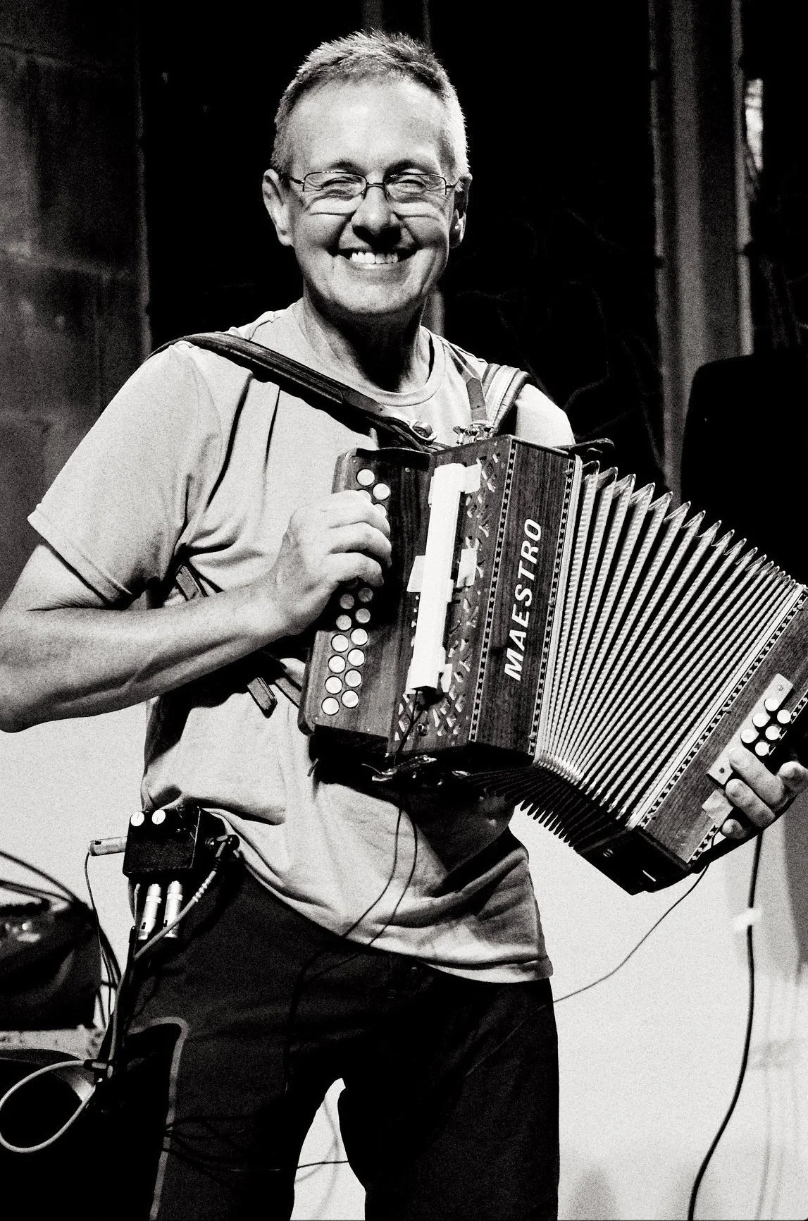A smiling person wearing glasses and a t-shirt, holding an accordion labeled 'MAESTRO' on a stage or performance setting.