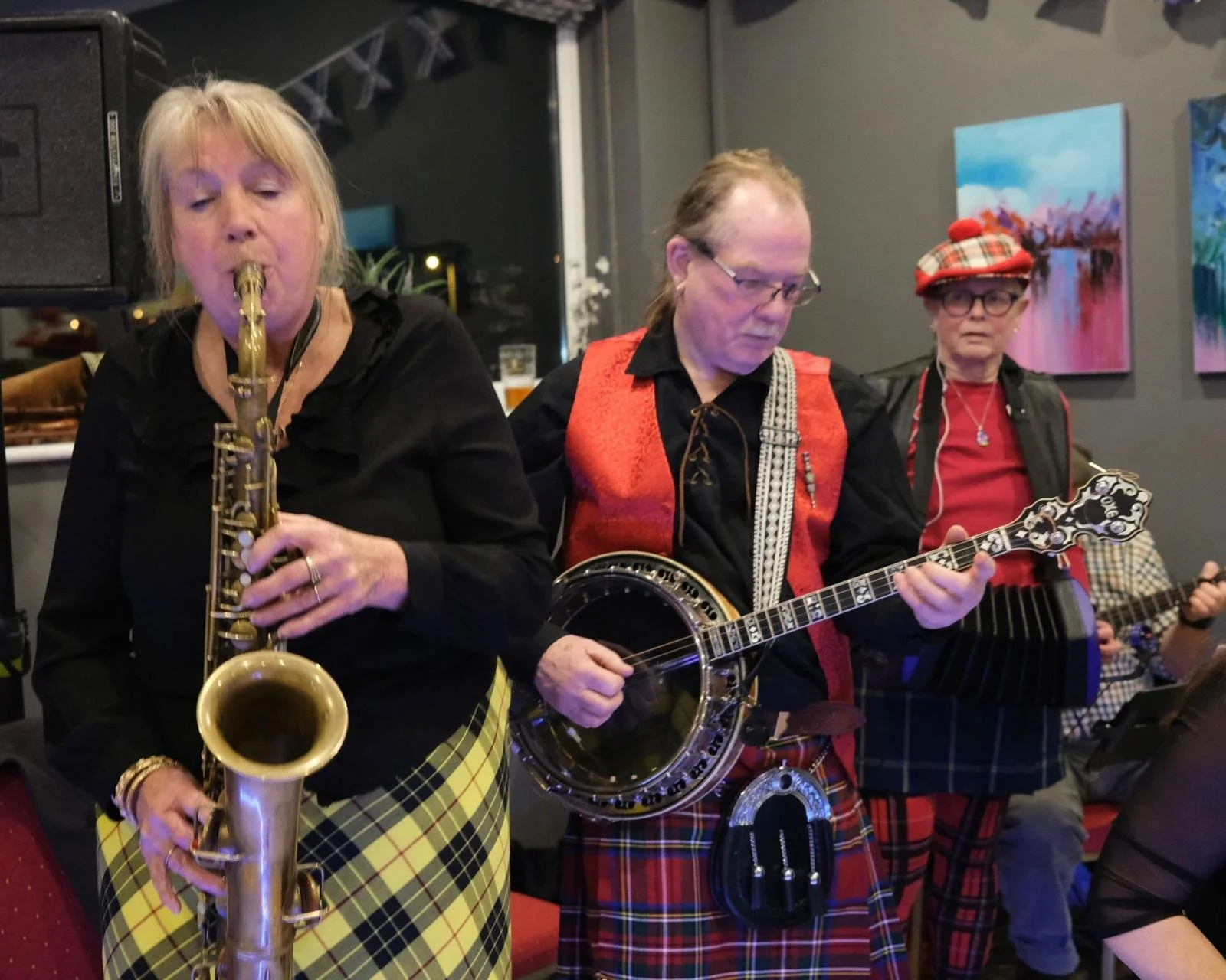 Three musicians playing instruments in a lively indoor setting. The woman on the left is playing a saxophone, while the man in the middle is playing a banjo, and the woman on the right is playing a guitar. All are dressed in colorful, patterned cloth