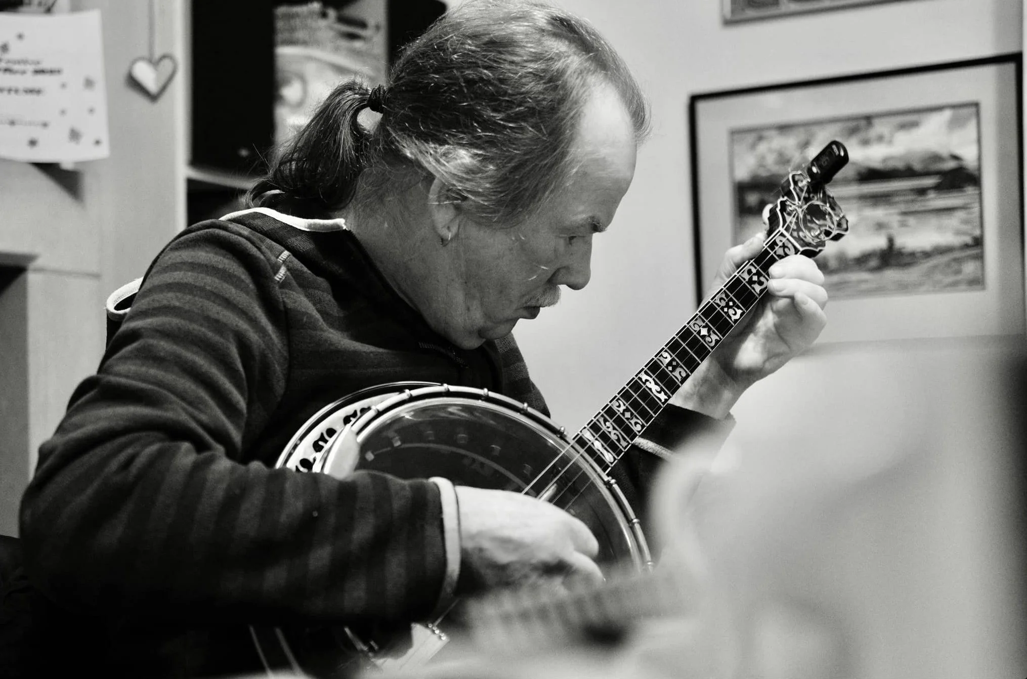 A man with long hair tied back playing a banjo, focused on his instrument, in a room with framed pictures on the wall.