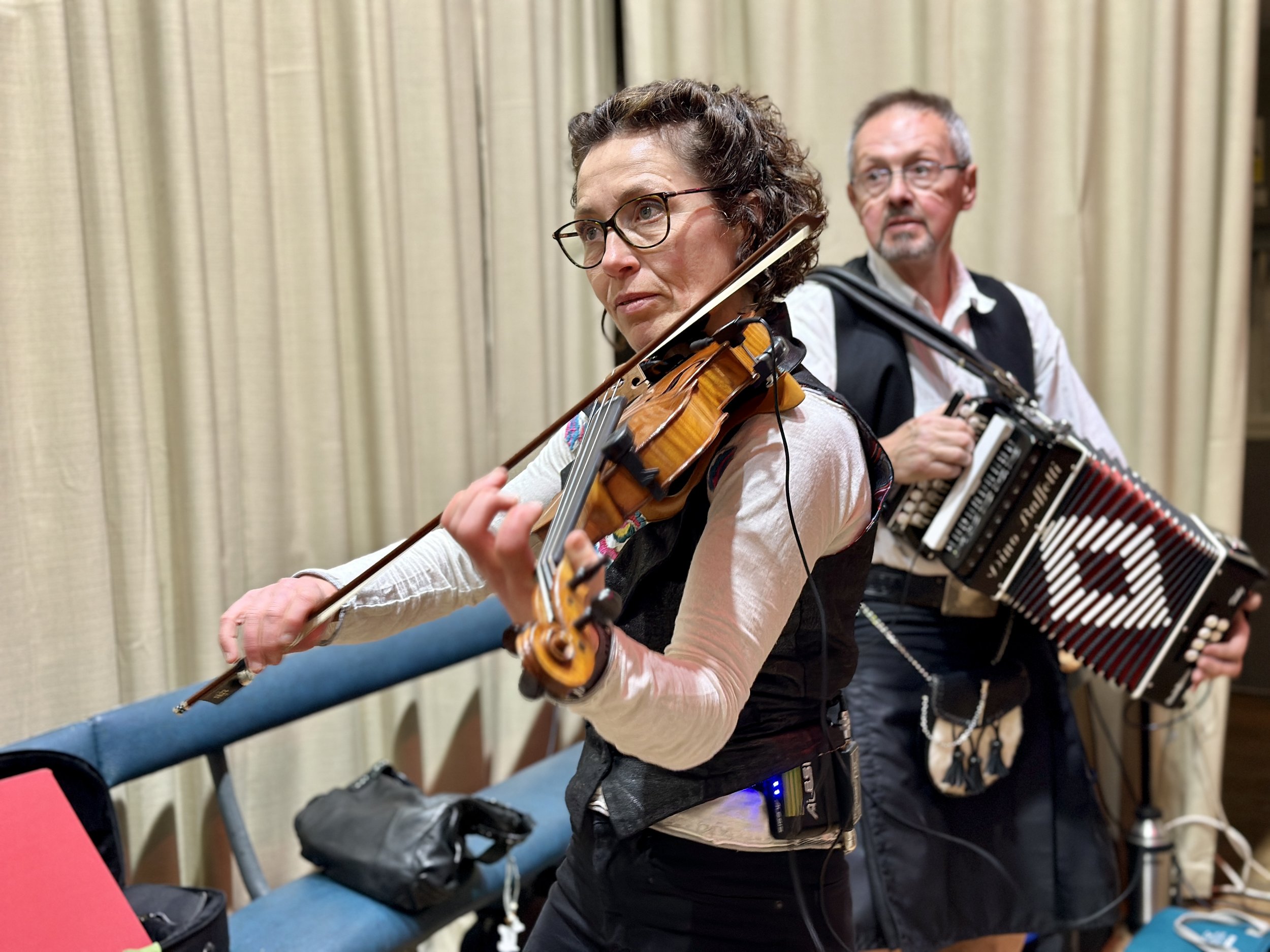 Two musicians, a woman playing a violin and a man playing an accordion, performing indoors with beige curtains in the background.