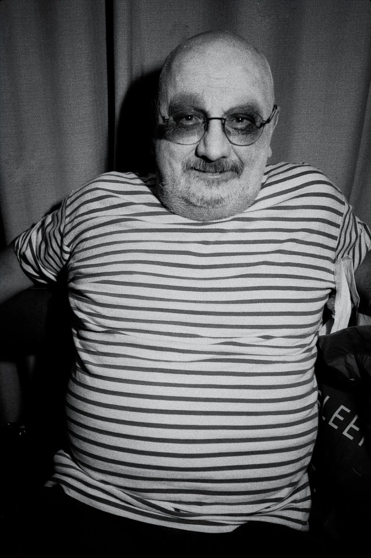Black and white photo of a smiling man with glasses and a beard, wearing a striped shirt, sitting in front of a curtain.