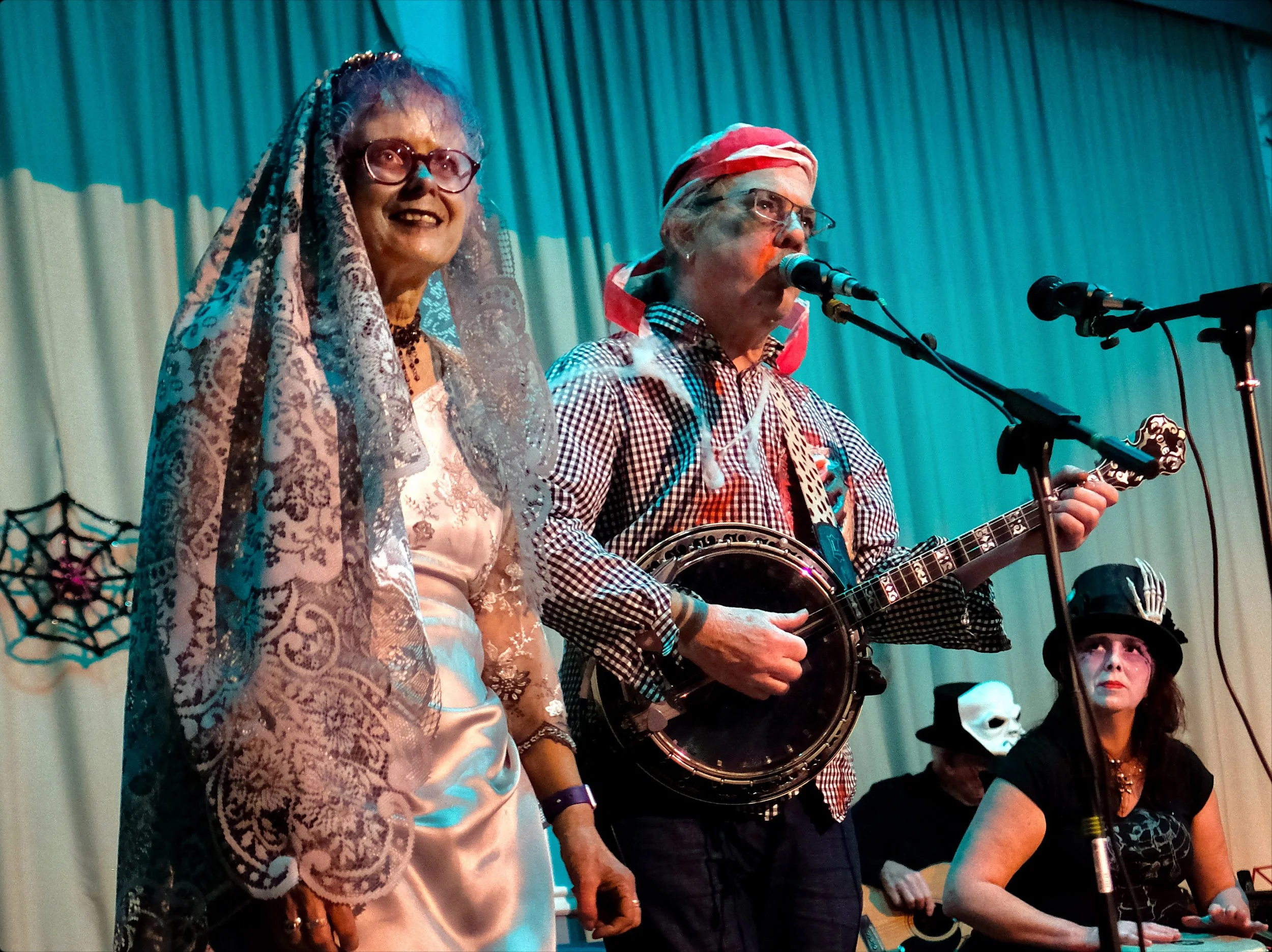 People performing on stage at a Halloween-themed event, with costumes including a woman in a lace shawl and satin dress, a man in checkered shirt with a tambourine and guitar, a woman in a black top hat with feathers, and others wearing masks, in fro