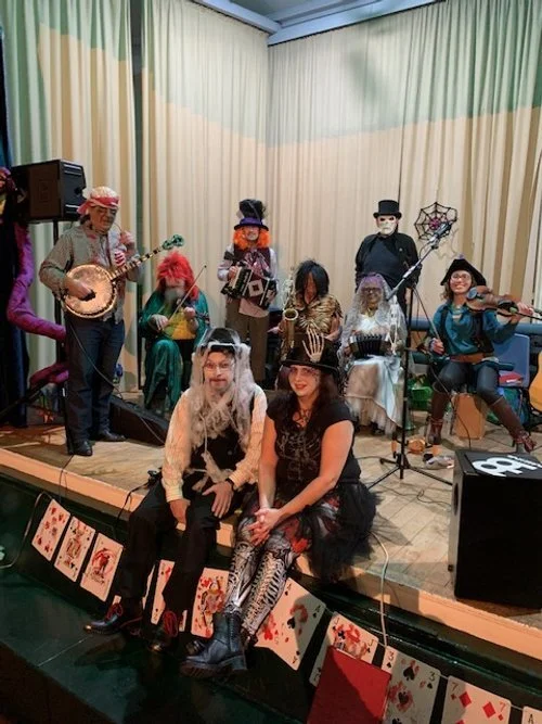 Children dressed in Halloween costumes performing on stage as a band, with two women in costumes sitting in front of the stage, and playing musical instruments in the background.