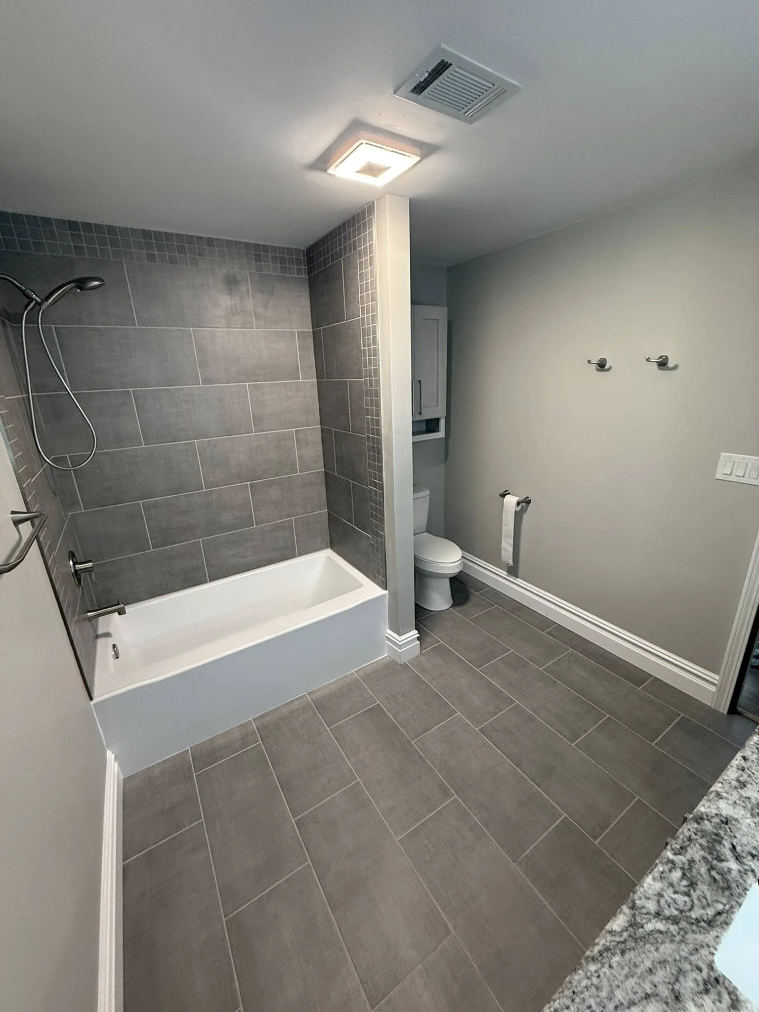 Modern bathroom with gray tiled shower area, a bathtub, and a toilet, with a towel bar and hooks on the wall.