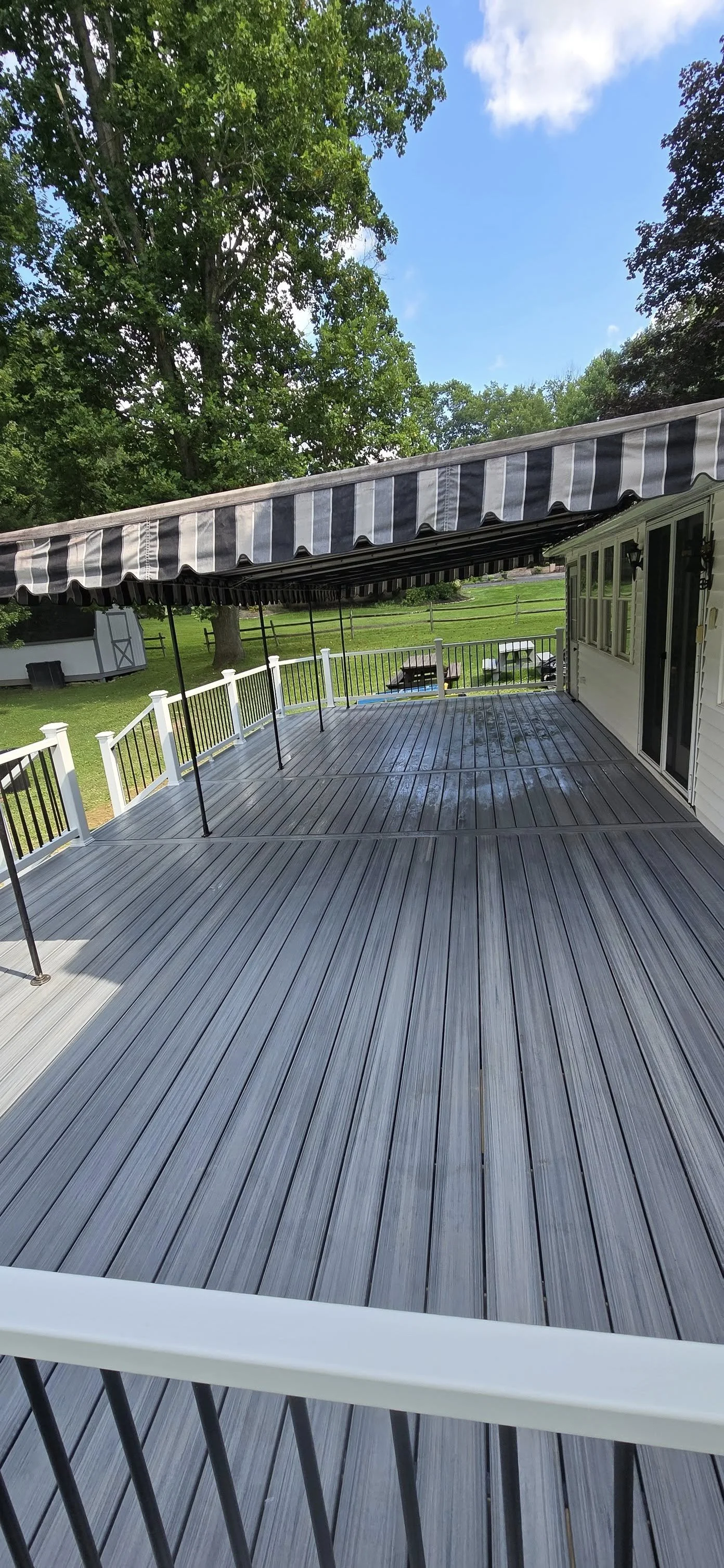 View of a large outdoor wooden deck with an arbor with black-and-white striped awning, surrounded by a white and black railing, overlooking a lush green backyard with trees, a shed, and picnic tables, under a partly cloudy sky.
