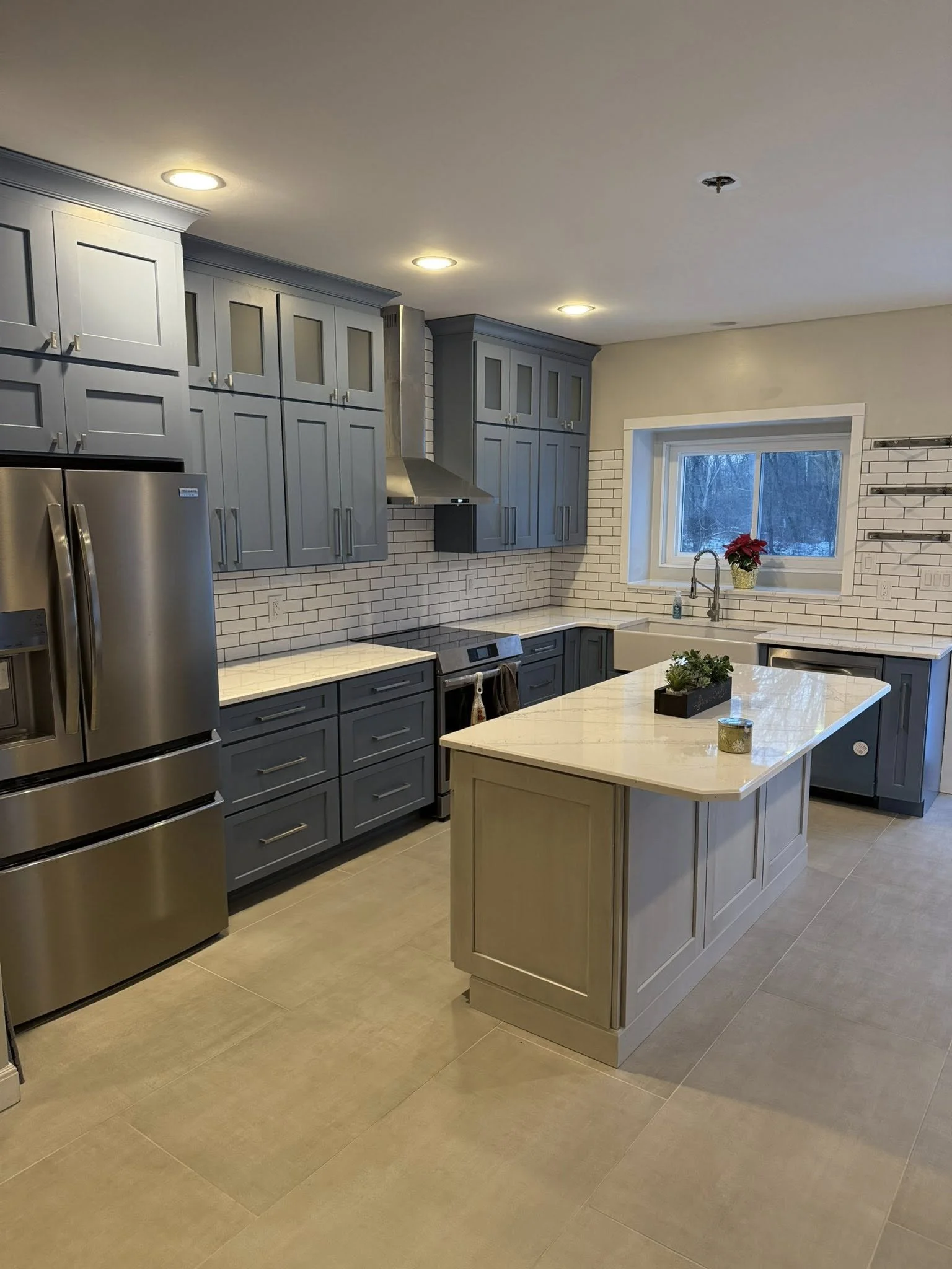 Modern kitchen with grey cabinets, white countertops, stainless steel appliances, and a white brick backsplash. An island with a light-colored countertop and a small potted plant on it. A window above the sink with a poinsettia plant, and some wall hooks on the right side.