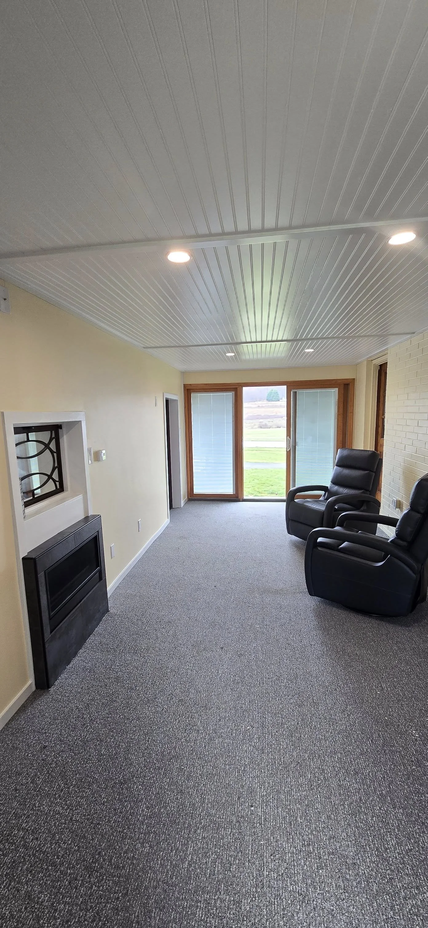 A cozy room with two black reclining chairs near a large sliding glass door, a fireplace with a decorative metal grille, and a view of a grassy outdoor area.