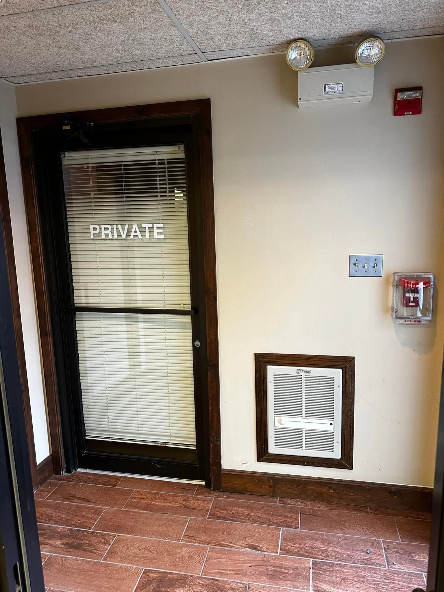 Interior of a building showing a glass door with a 'PRIVATE' sign, a wooden floor, and wall-mounted safety equipment including a fire alarm pull station, a fire sprinkler, and an air vent.