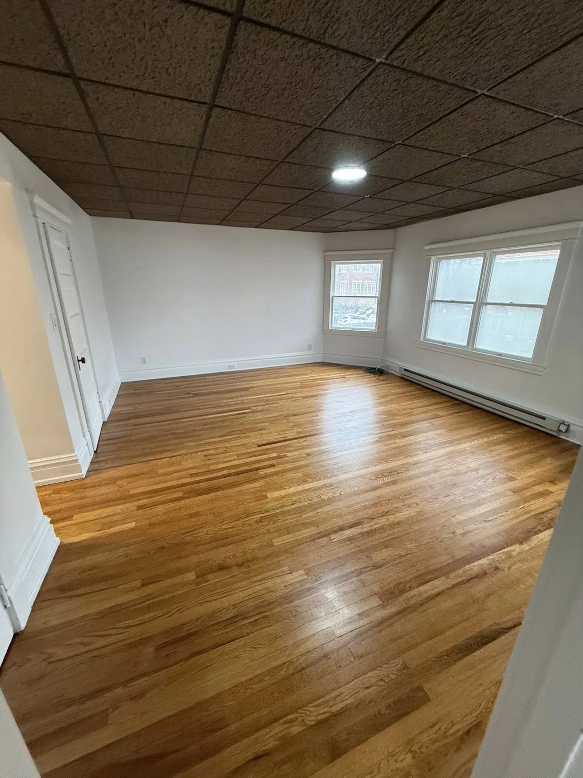 Empty living room with wood flooring, black and brick fireplace, wooden mantel, and minimal furniture, illuminated by ceiling lights.