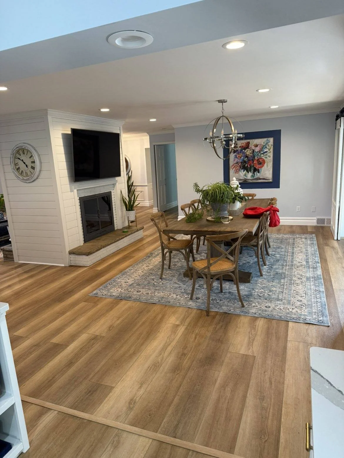 A cozy dining room with a wooden table and eight chairs on a blue patterned rug, a pendant light fixture, a framed floral painting on a light gray wall, a white fireplace with a large flat-screen TV above, and a fireplace with a clock on the wall. There's a potted plant on the table, and sliding glass doors with vertical blinds.