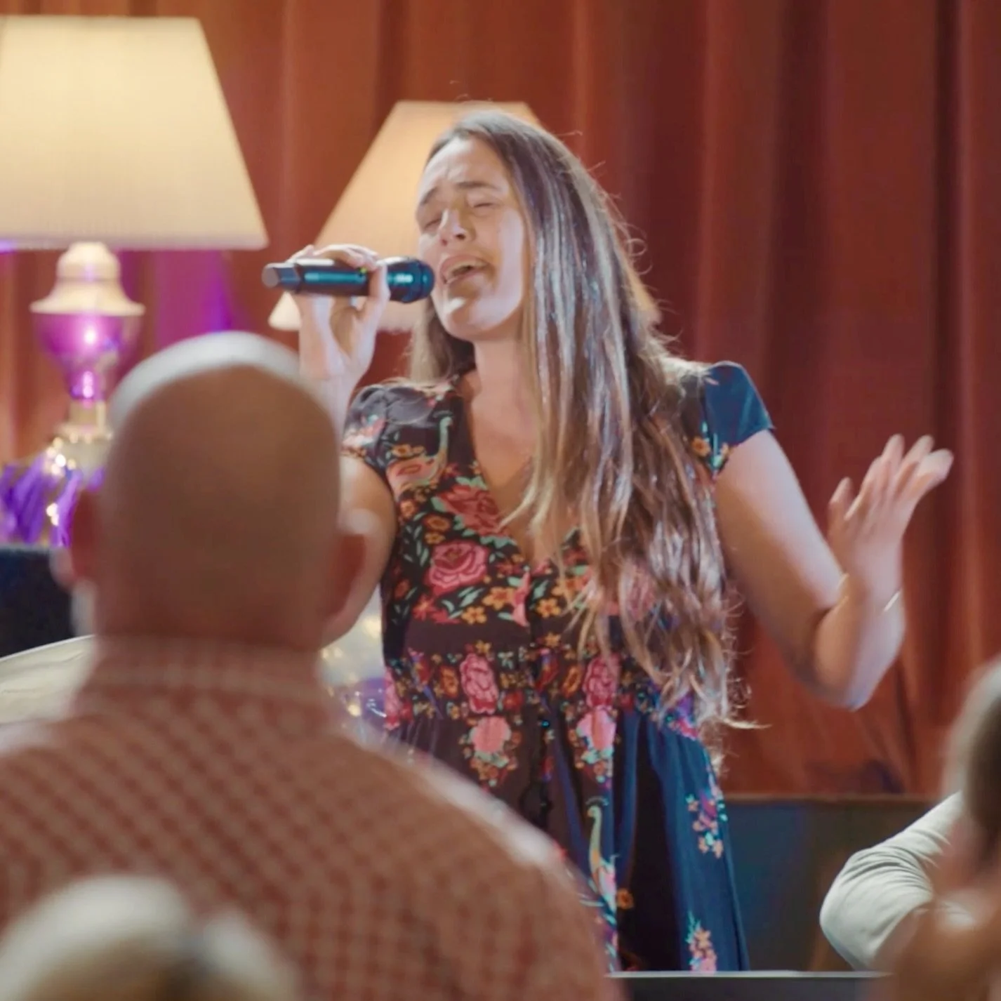 A woman with long brown hair singing into a microphone in a decorated indoor setting, with a red curtain in the background and people in the foreground.